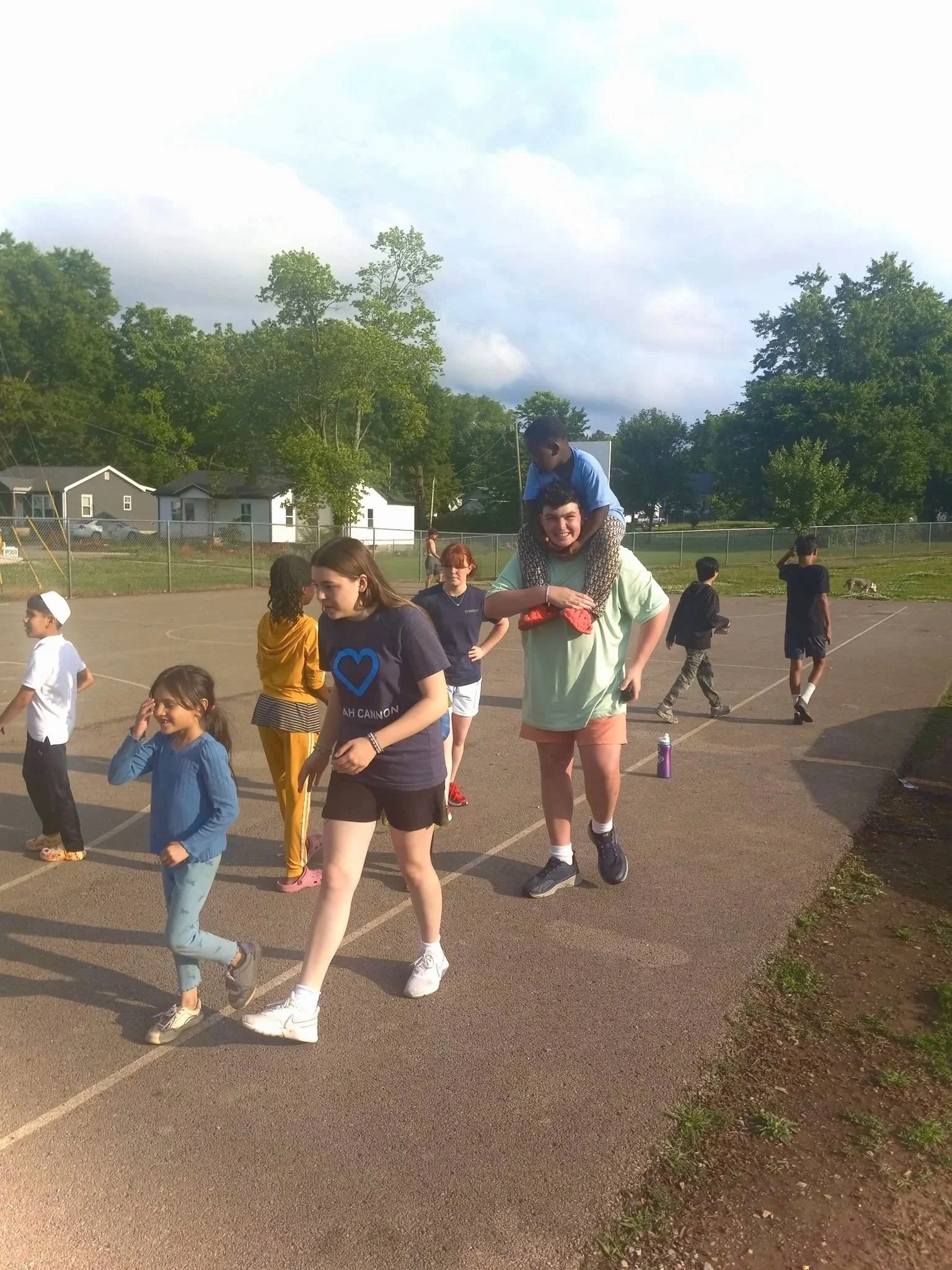 Children playing and walking on a school outdoor sports court with a chain-link fence, with trees and houses in the background, in a sunny setting.
