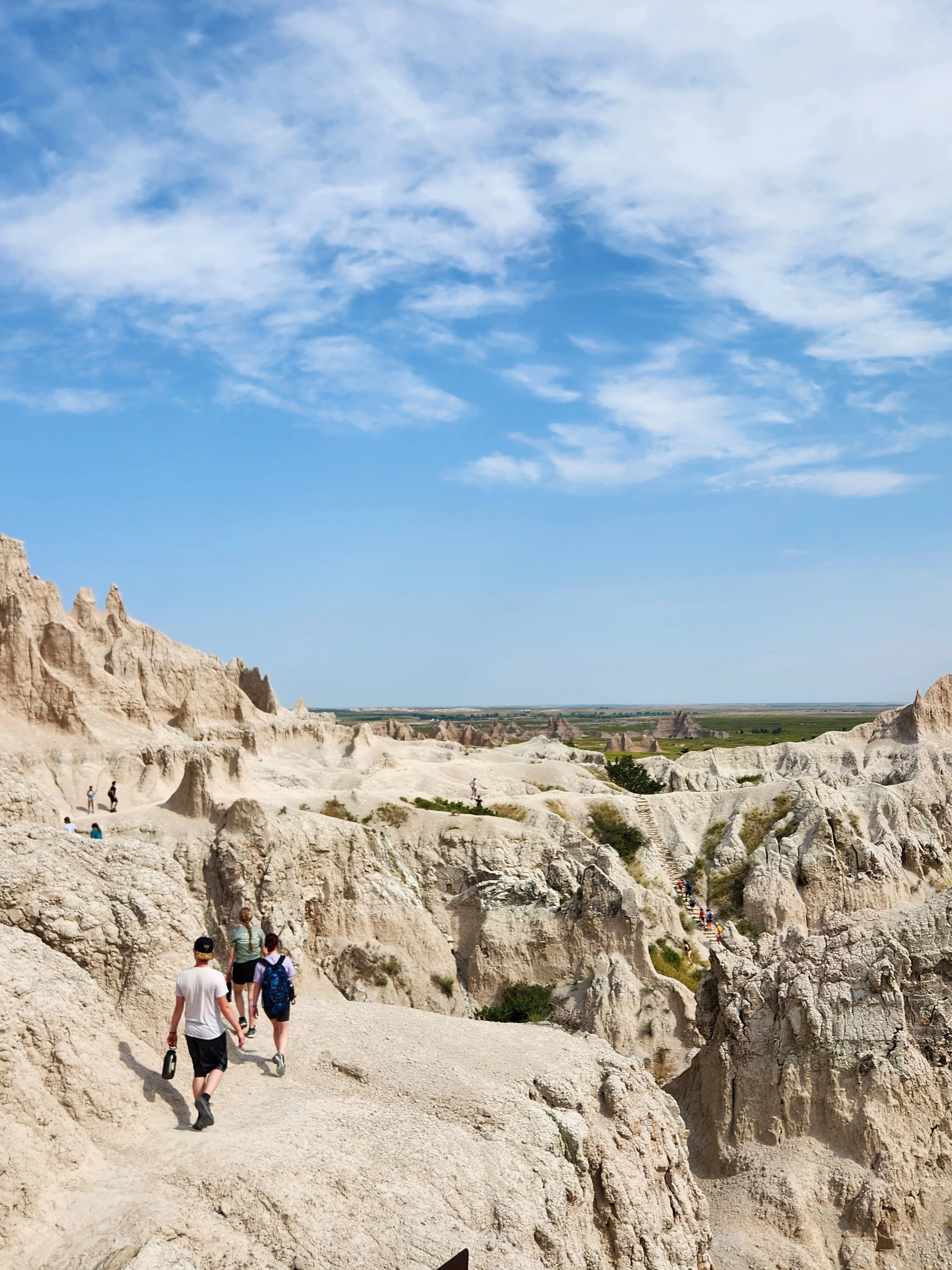 People hiking along a trail through a dry, rocky, desert-like landscape with eroded rock formations under a blue sky with scattered clouds.