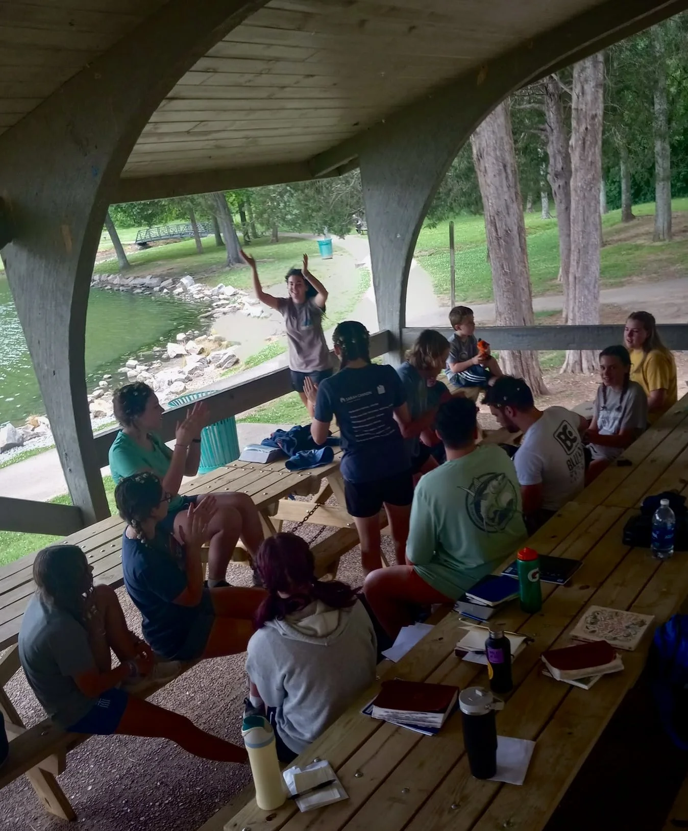 Group of people gathered under a shelter by a lake, participating in a group activity or discussion, with trees and a walking path in the background.