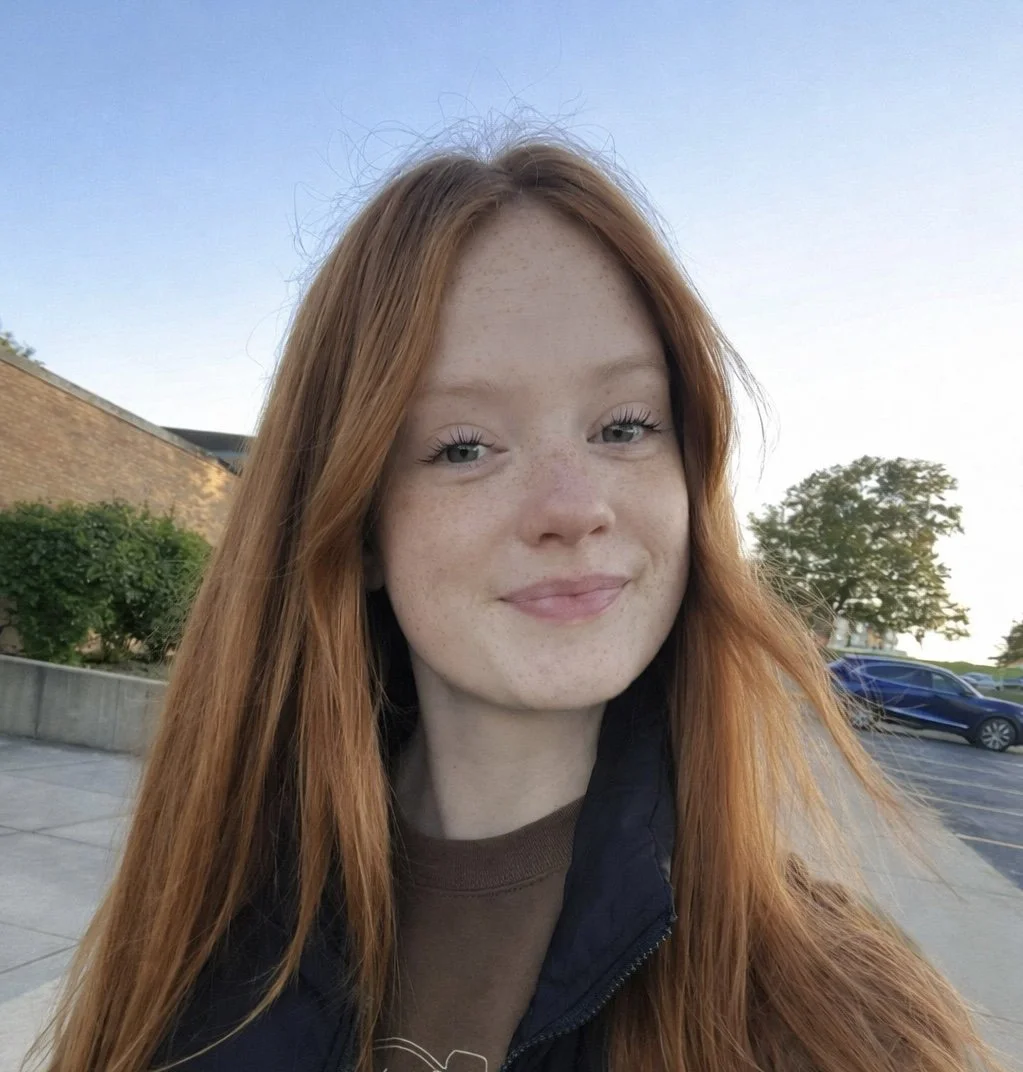 A red-haired girl smiling and holding a black and white goat kid outdoors on a sunny day, with a wooden fence, green grass, and trees in the background.