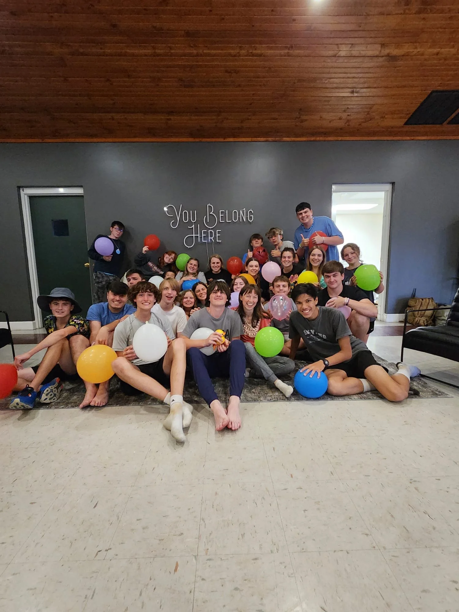 Group of teenagers and young adults sitting on the floor with colorful balloons, smiling for a photo, with a wall behind them that says "You Belong Here".