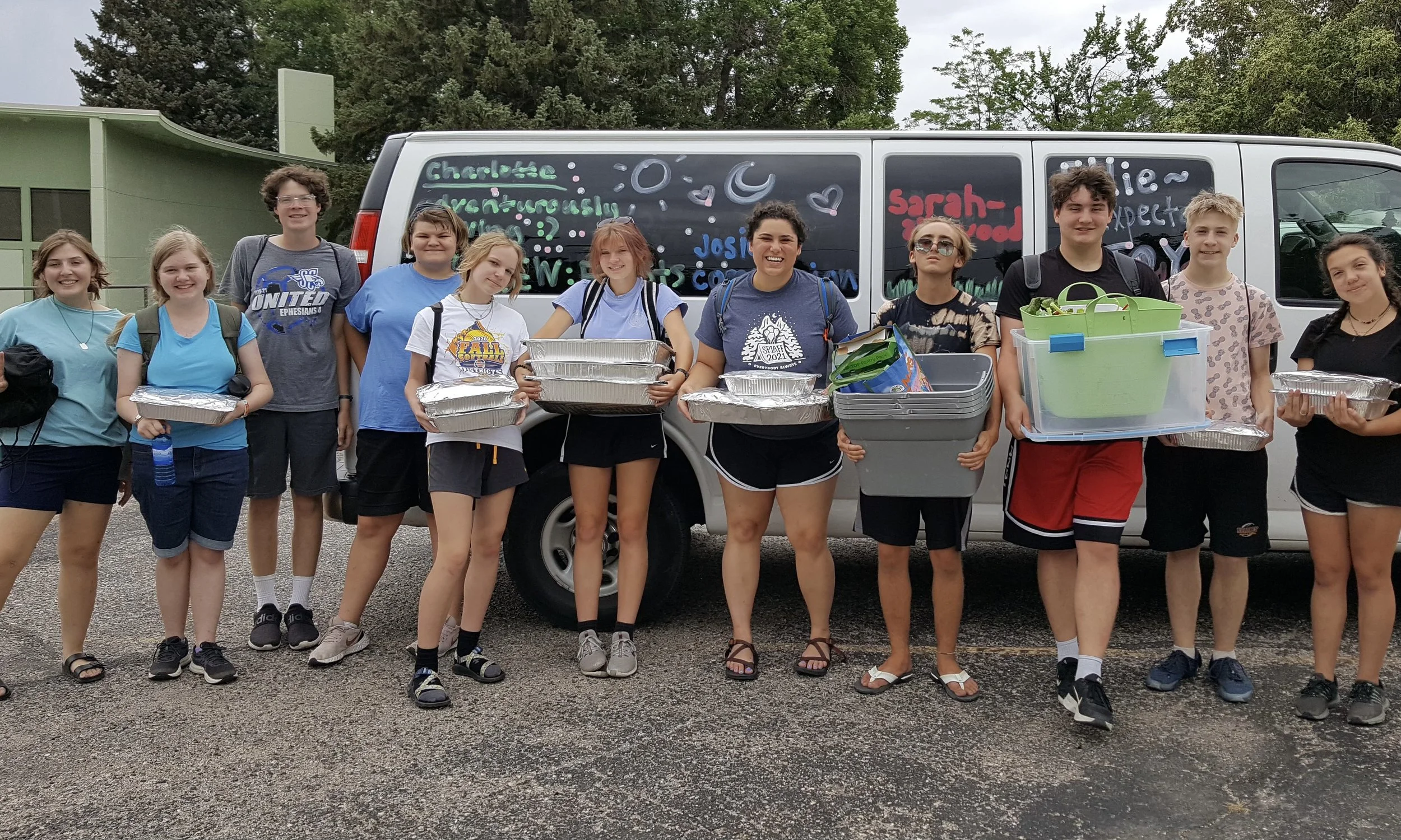 Group of ten teenagers standing in front of a van with colorful handwritten messages, holding lunch trays and supplies, outdoors on a cloudy day.