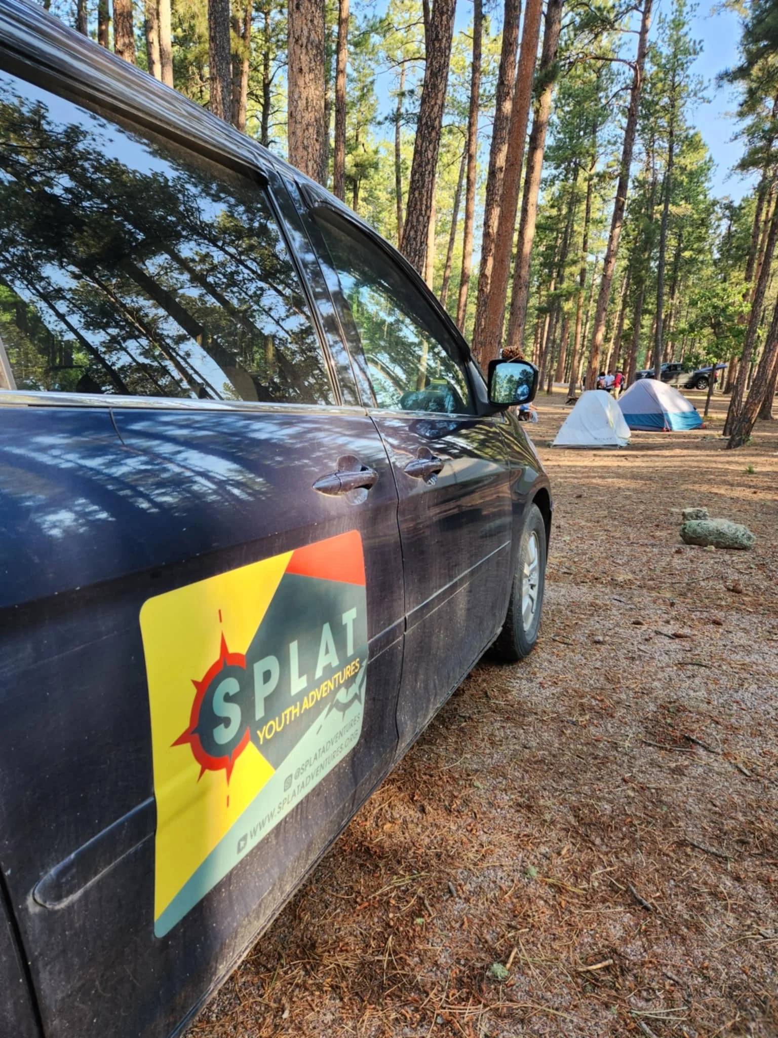 A black SUV with a SPLAT Youth Adventures logo parked on a forest campground with tents in the background, tall pine trees, and a dirt ground.