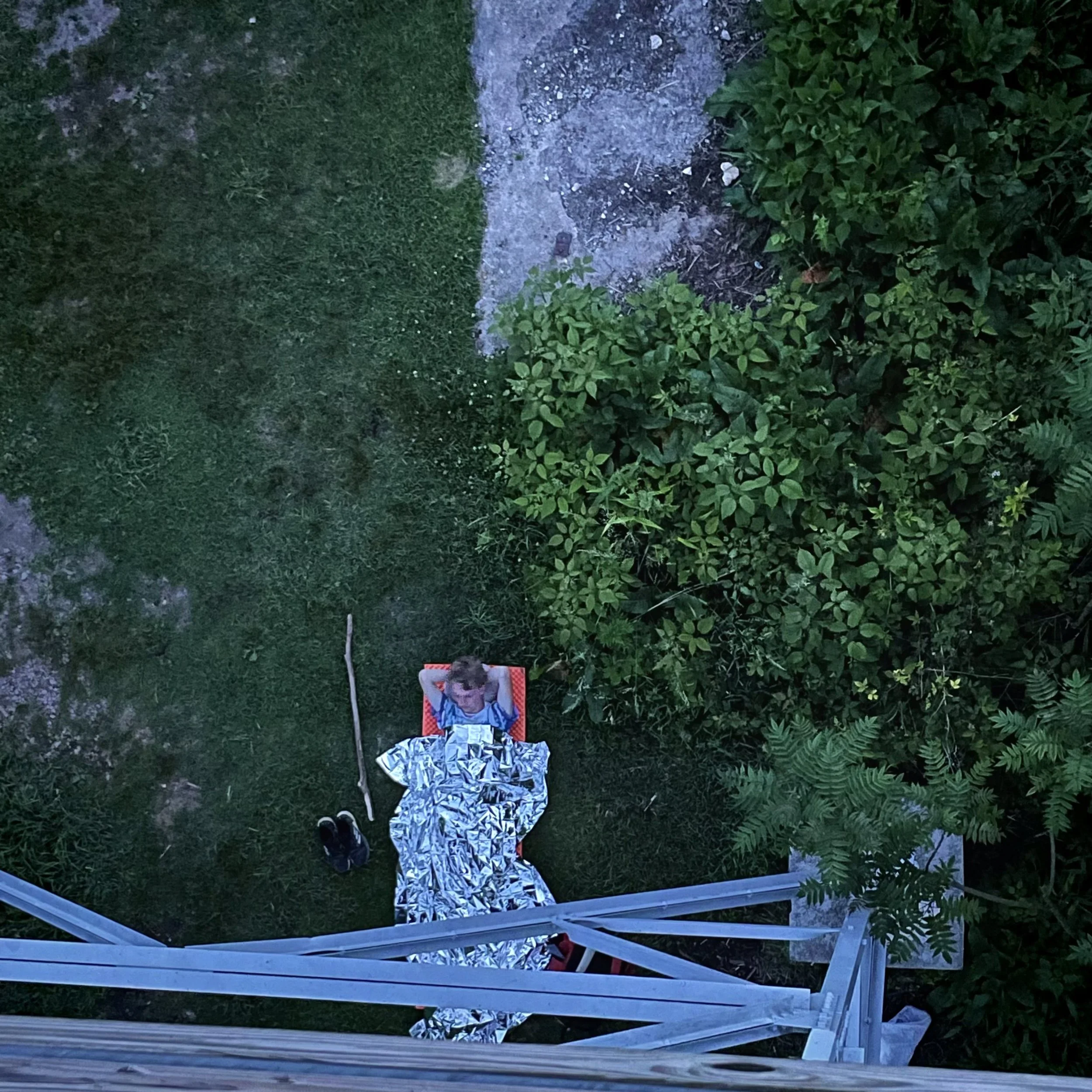 A person is lying on a red lounge chair covered with a patterned blanket in a backyard, resting with arms behind their head. Shoes are placed on the grass nearby, surrounded by green plants and trees.