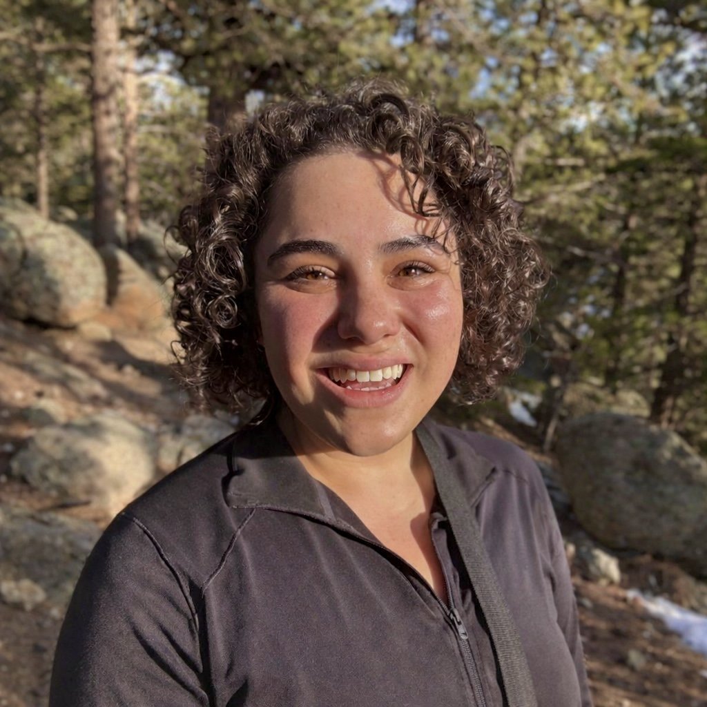 A woman with curly hair smiling outdoors in a forest with trees and rocks.