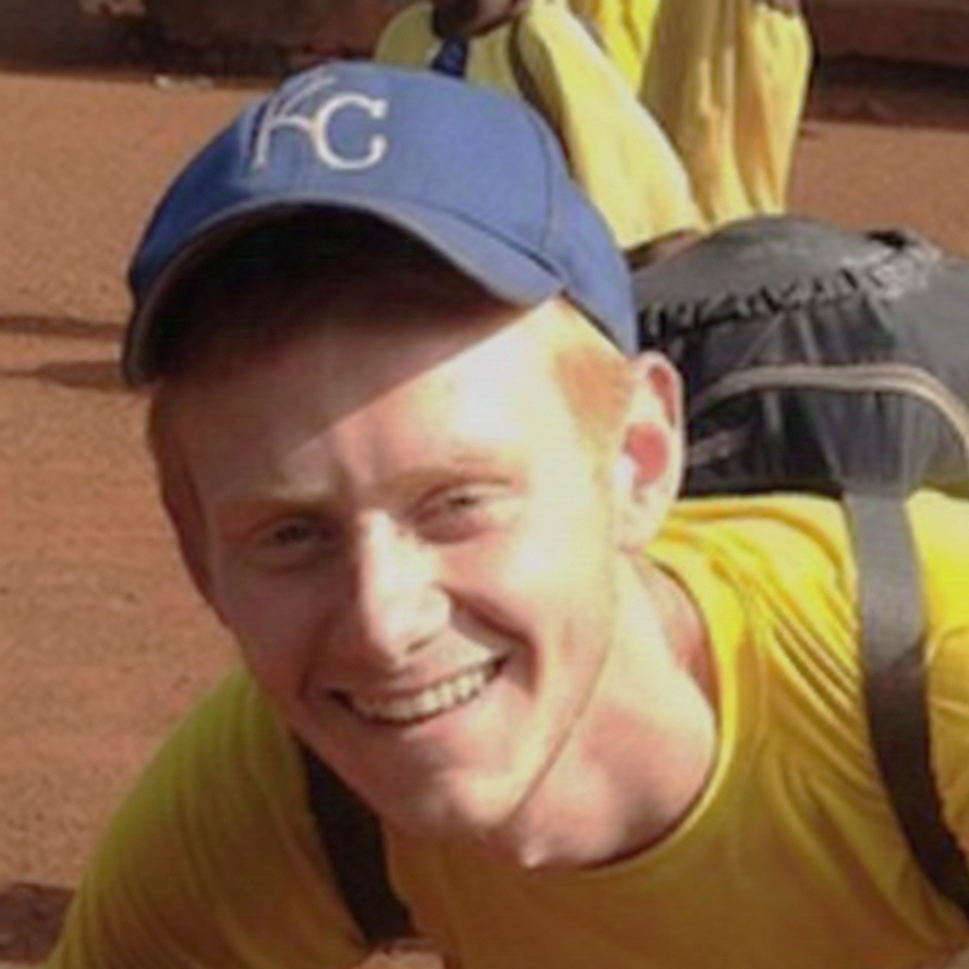 A young man smiling, wearing a blue baseball cap with white letters 'KC', a yellow shirt, and carrying a backpack, outdoors on a clay or dirt surface.