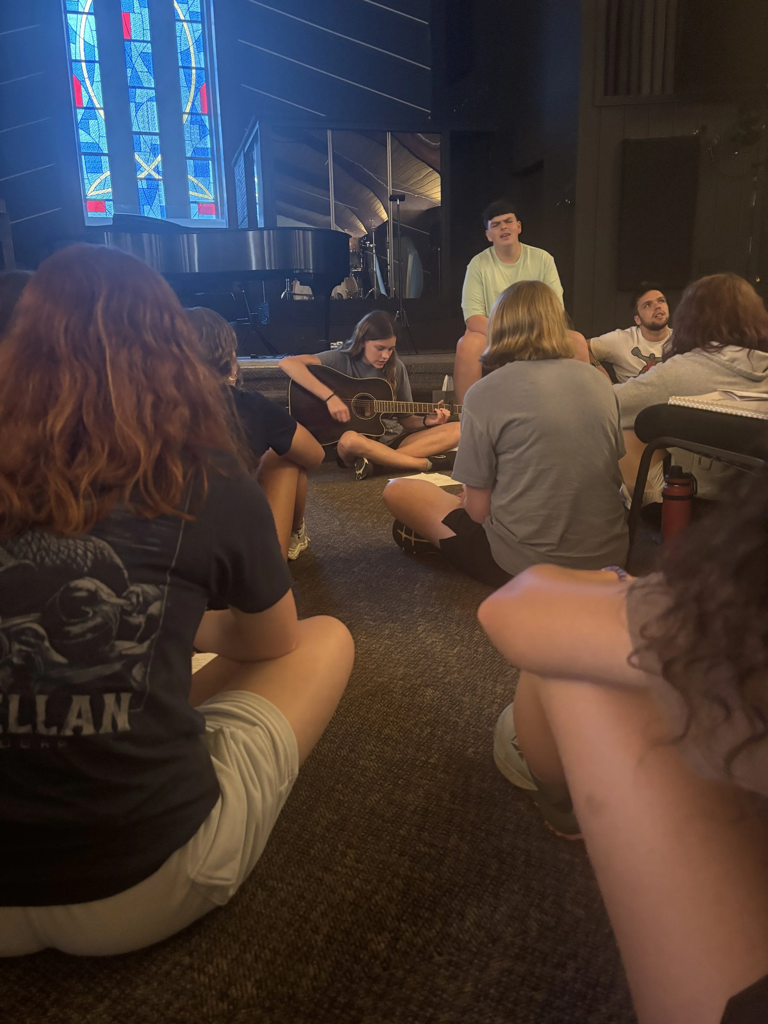 Group of young people sitting on the floor in a circle, with one person playing a guitar and another singing, during a casual music session in a church or music hall with stained glass windows.