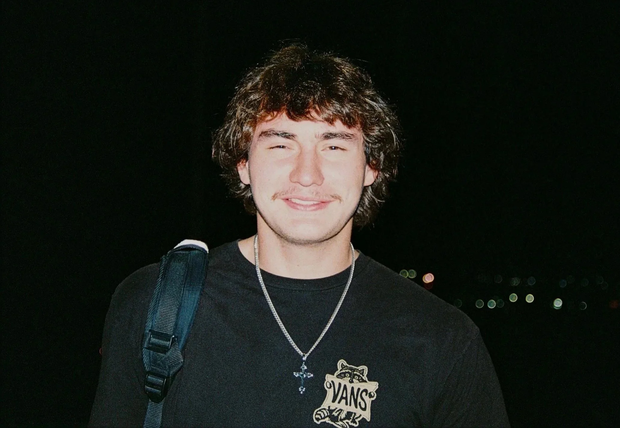 A young man with brown, wavy medium-length hair, smiling, wearing a black Vans t-shirt with a small graphic at the chest, a silver necklace with a cross and blue pendant, and carries a black backpack on his shoulder, standing outdoors at night with distant lights in the background.