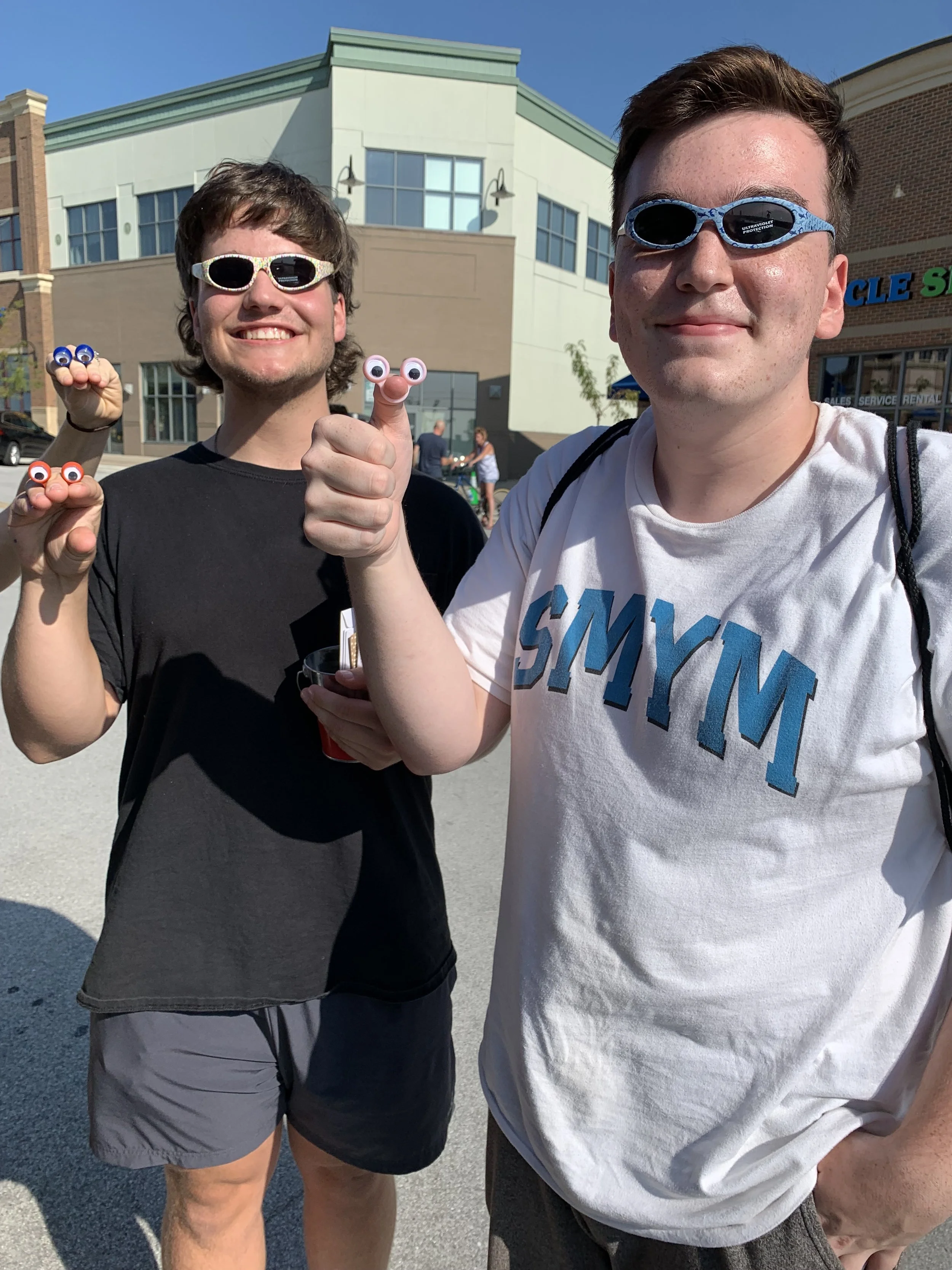 Two young men outdoors wearing sunglasses, smiling, and showing finger puppets with googly eyes, with a shopping strip mall in the background.
