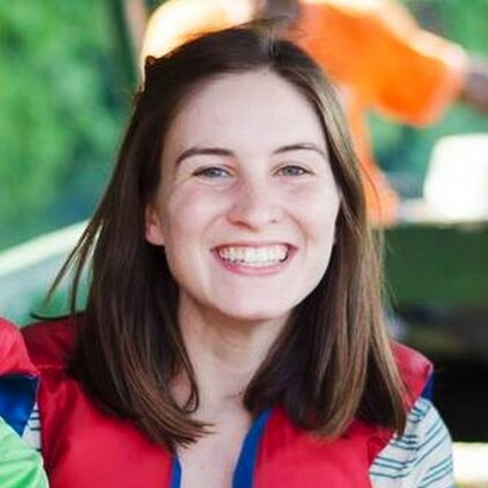 Close-up of a smiling young woman with shoulder-length brown hair, wearing a red jacket with blue and striped details, indoors with a blurred background.