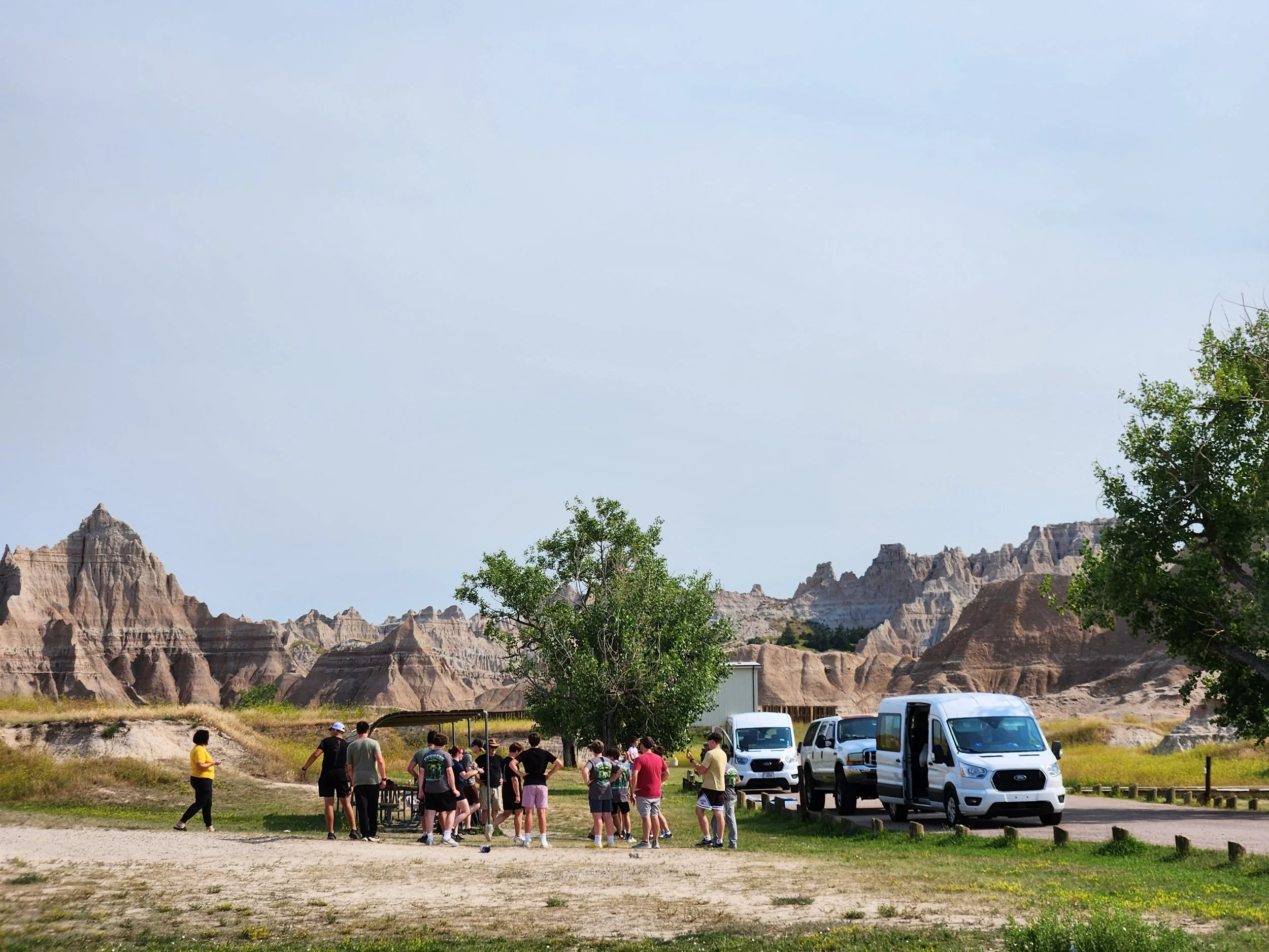 Group of people at a tour shuttle stop in Badlands National Park, South Dakota, with rugged eroded rock formations and a clear sky in the background.