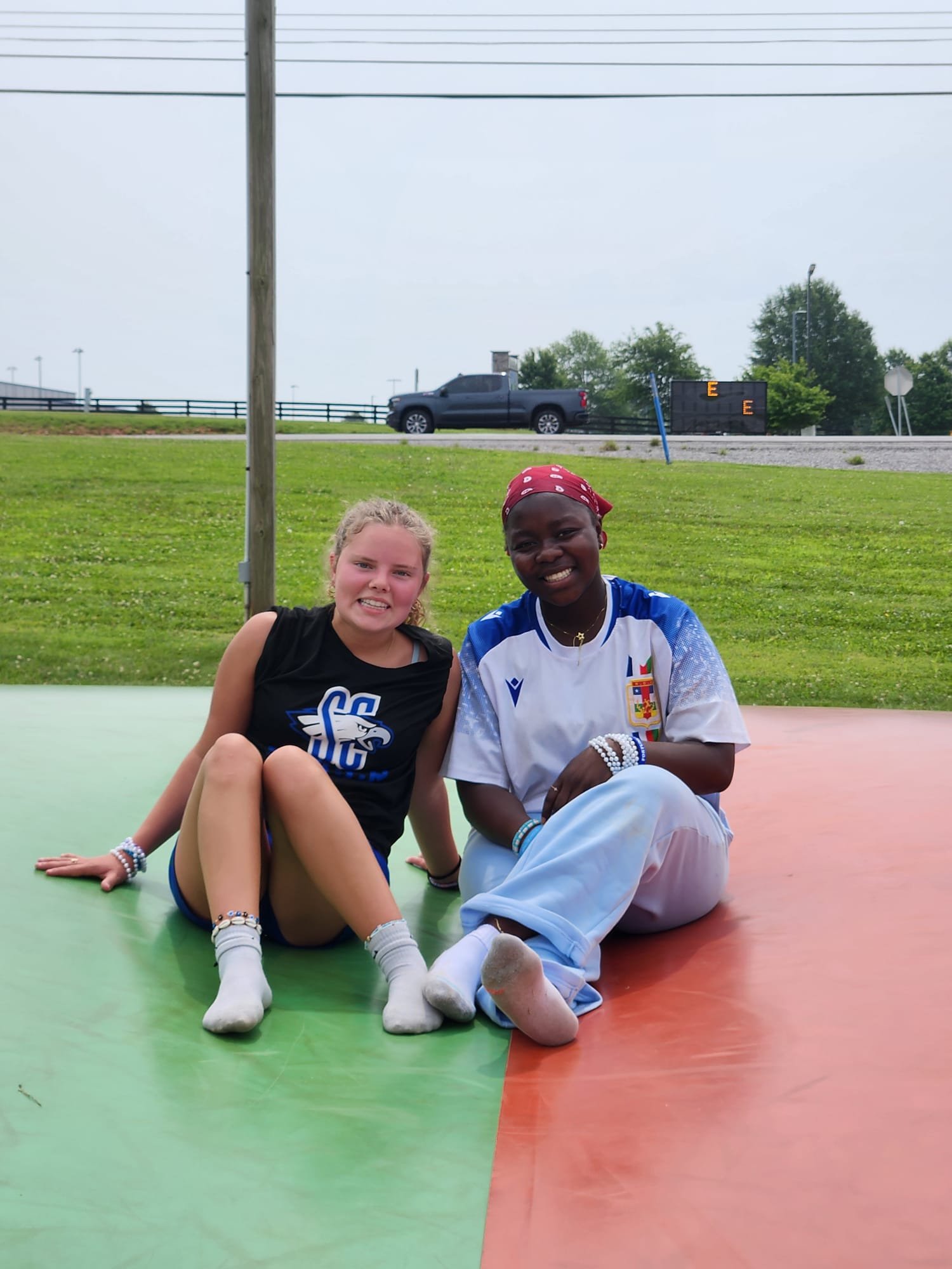 Two young women, one with blonde hair and the other with dark skin, sitting on a colorful outdoor mat, smiling at the camera, with a grassy field and a black truck in the background.