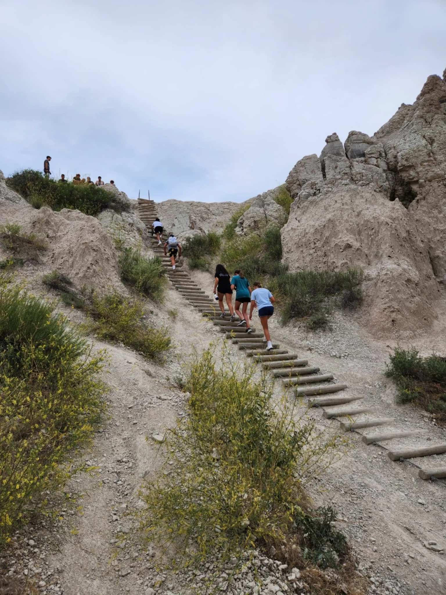 People hiking up a rocky, dirt trail with wooden steps in a mountainous terrain under cloudy sky.