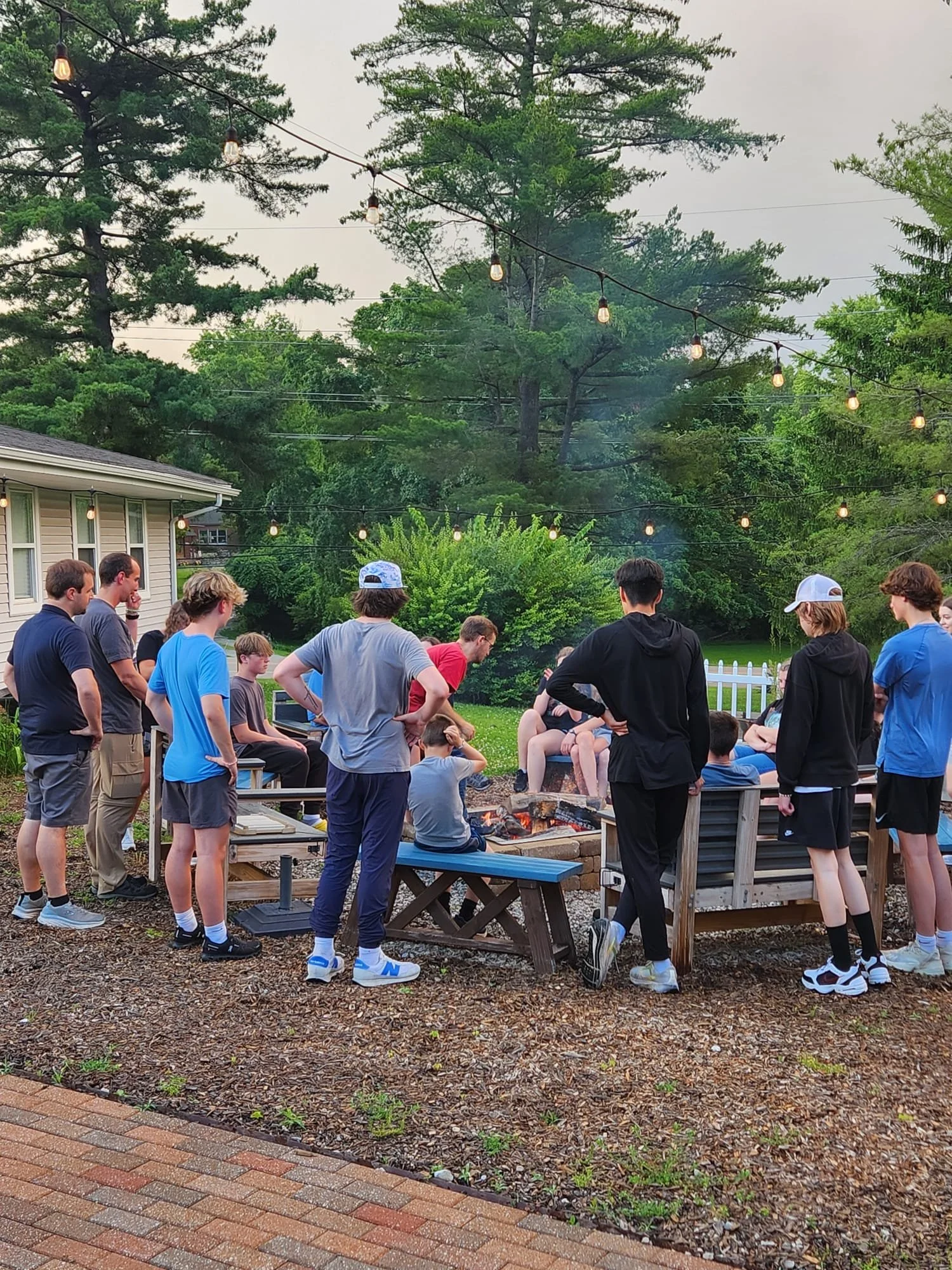 A group of young people gathered around a backyard fire pit in the evening, watching a fire while enjoying a social gathering outdoors under string lights.