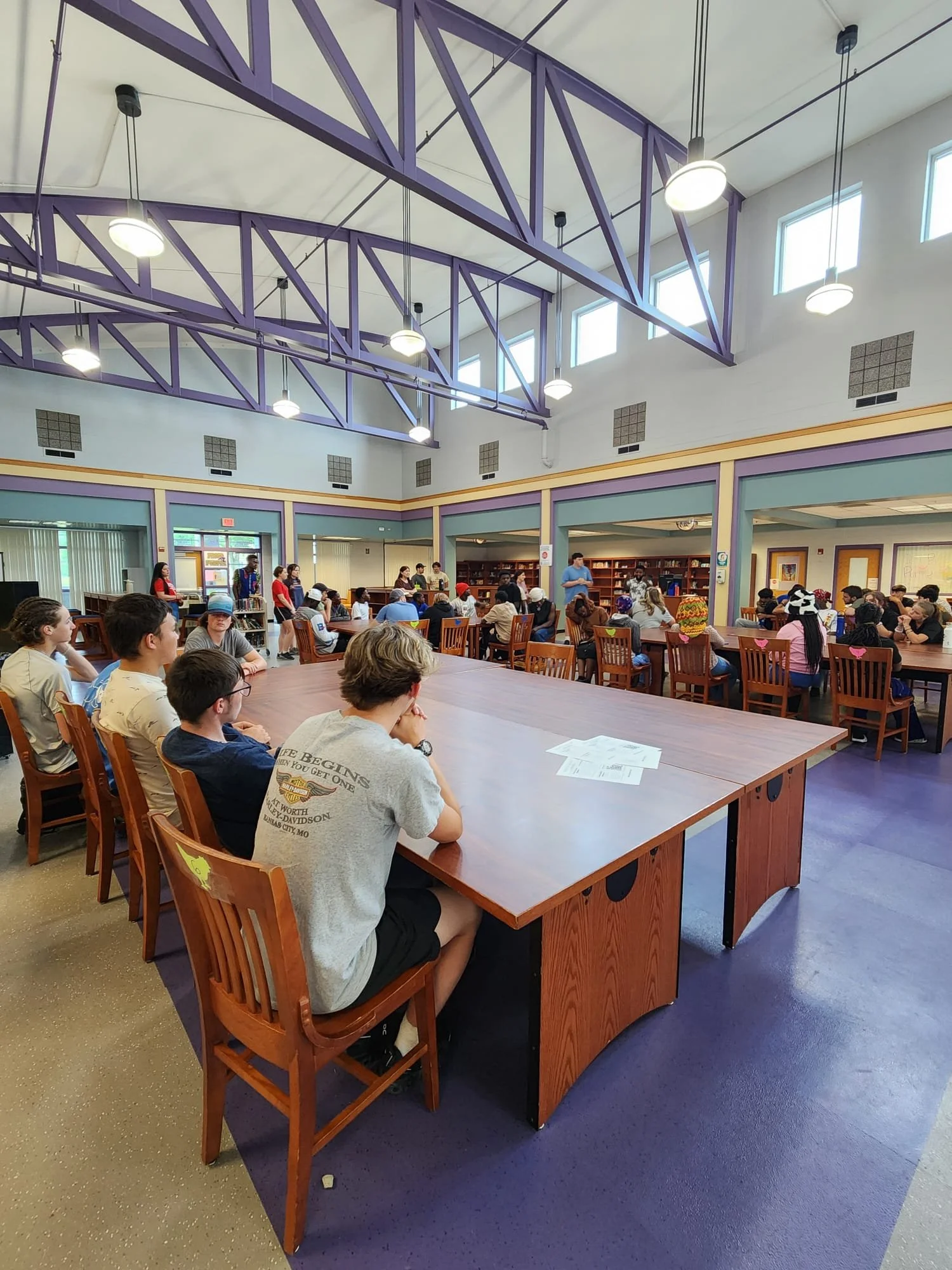 A large group of people seated at tables inside a colorful library or community center with high ceilings, purple beams, and large windows.