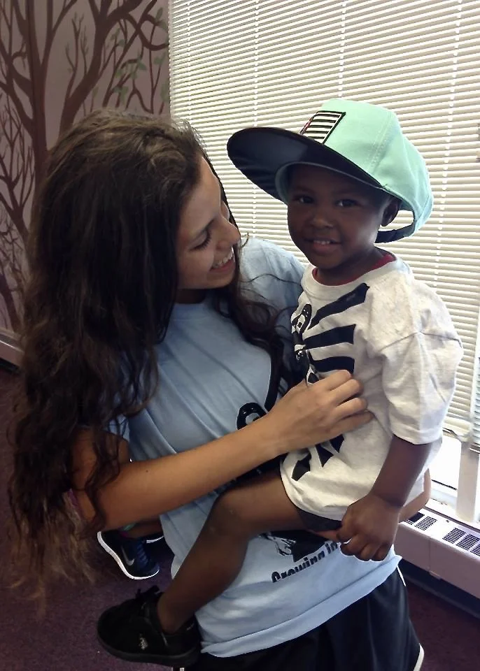 A woman smiling and holding a young boy with a baseball cap and striped shirt, near window with blinds, in a room with patterned wallpaper.