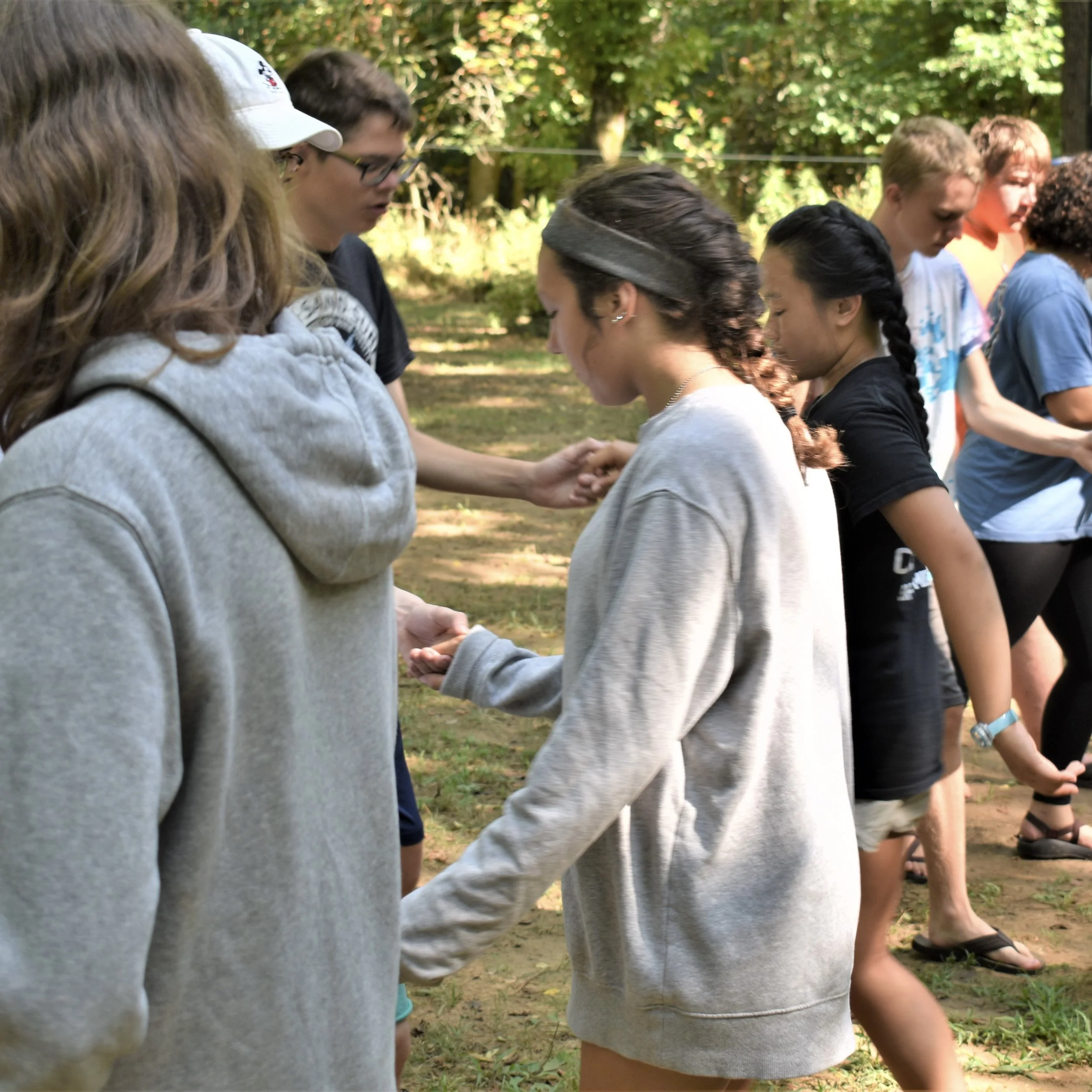 Group of young people standing in a line outdoors, holding hands in a circle on a grassy area with trees in the background.