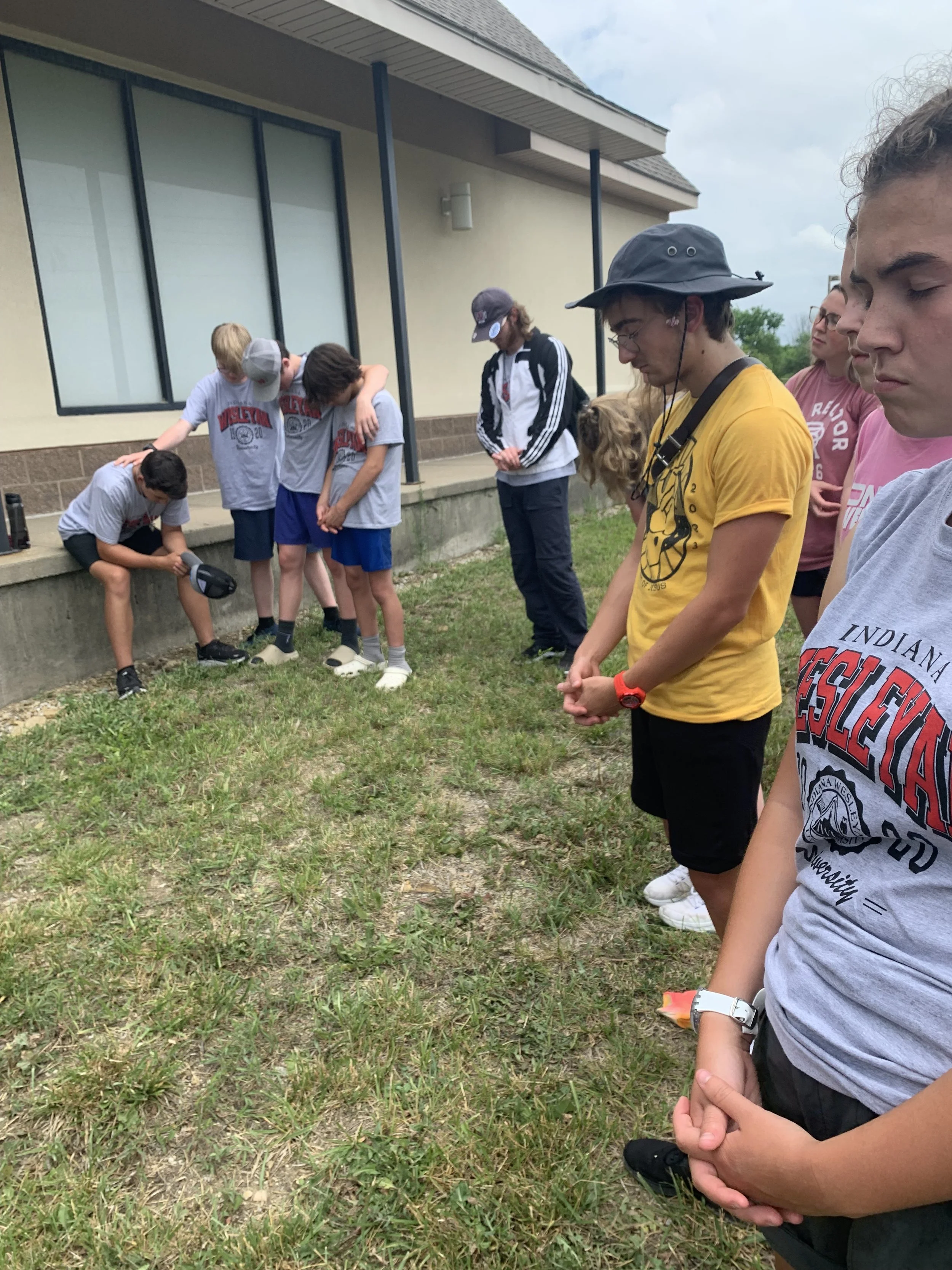 Group of children and teenagers standing outdoors near a building, with some bowing their heads in prayer or reflection. The children are wearing casual clothes, and some are in school uniforms. The setting appears to be a school or community area.