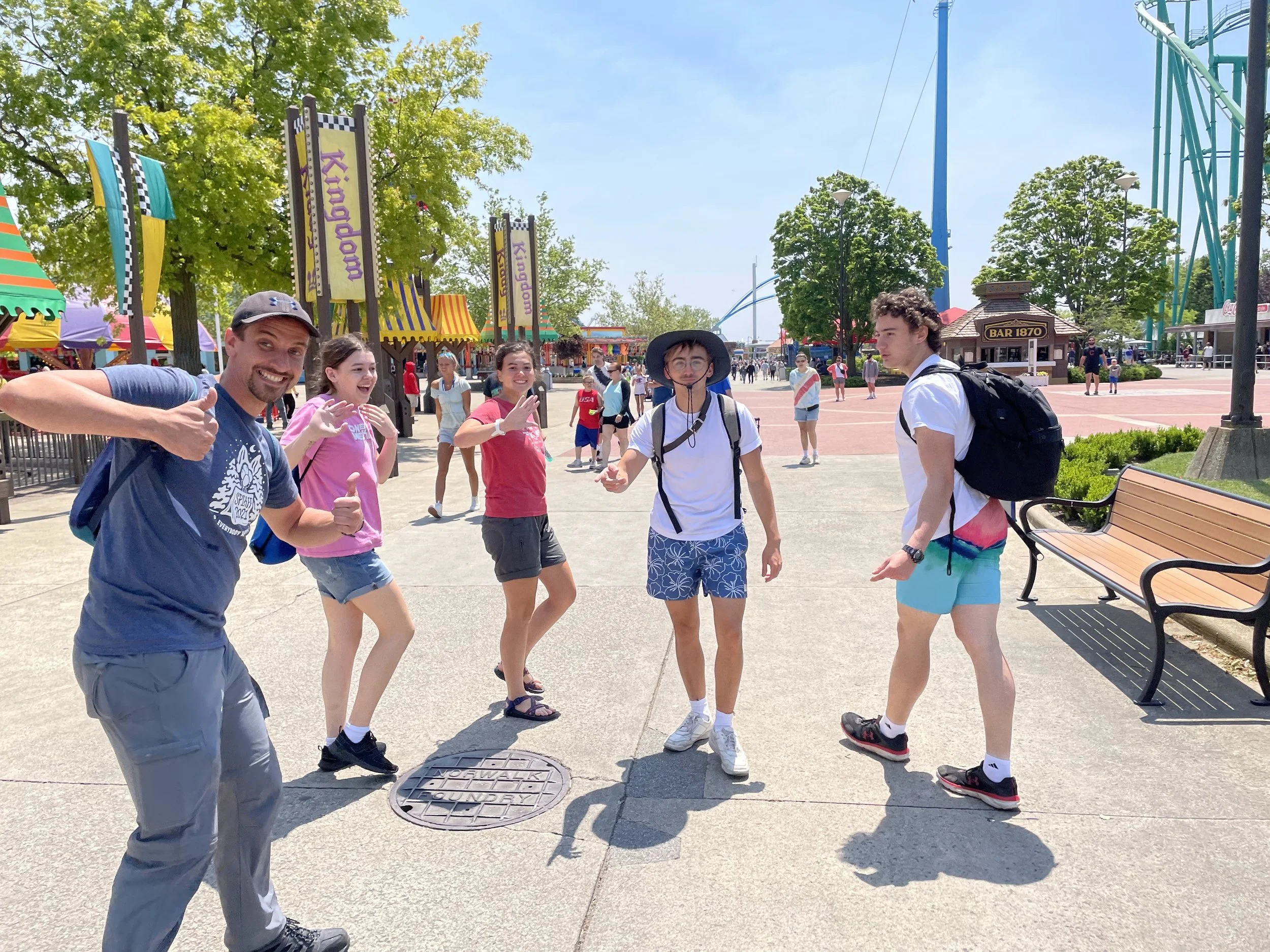 Group of five friends at an amusement park, posing for the camera with smiles and thumbs up, colorful rides and trees in the background on a sunny day.