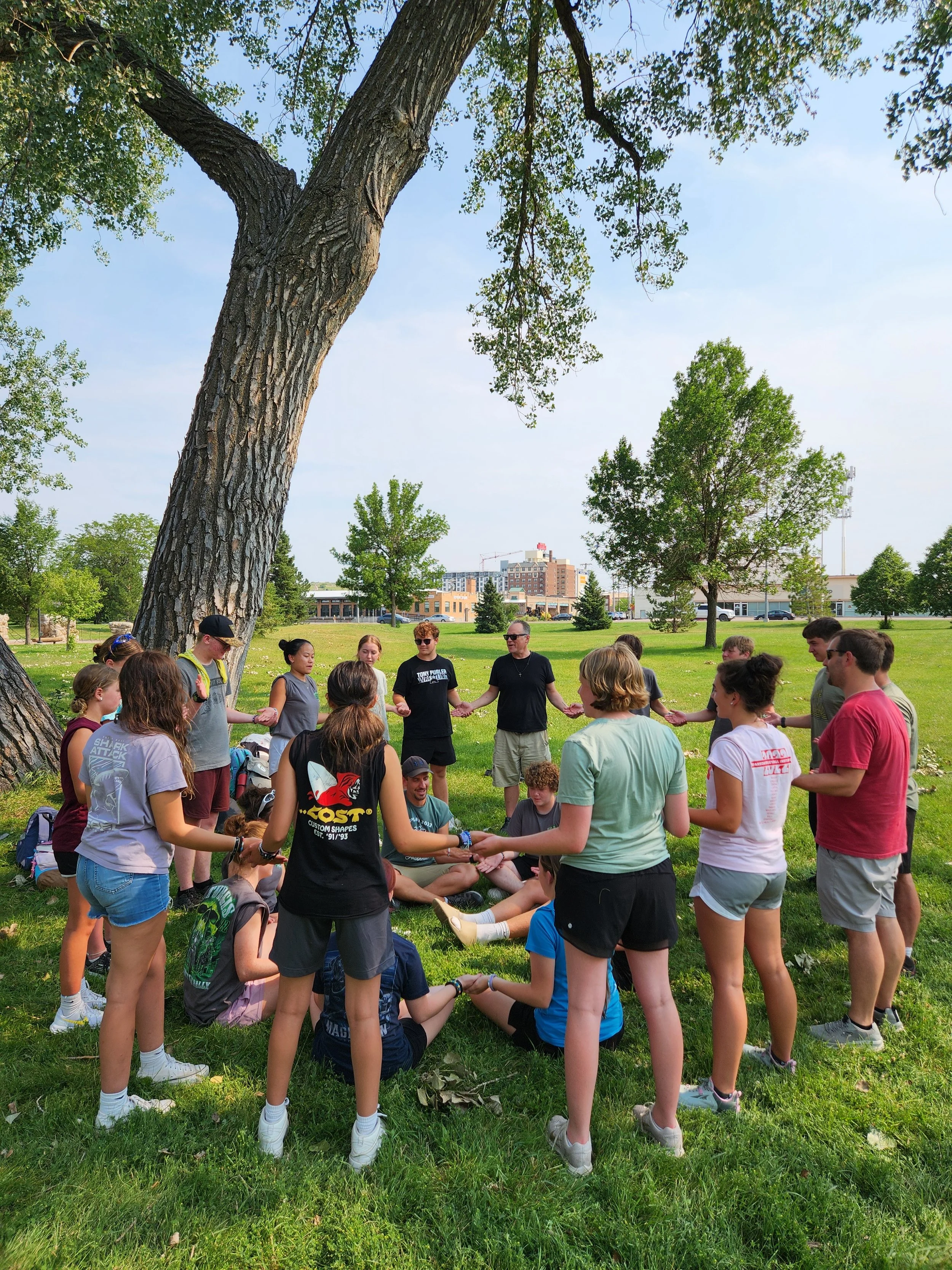 A group of children and teenagers holding hands forming a circle outdoors in a park, with some seated on the grass and others standing, under a large tree on a sunny day.