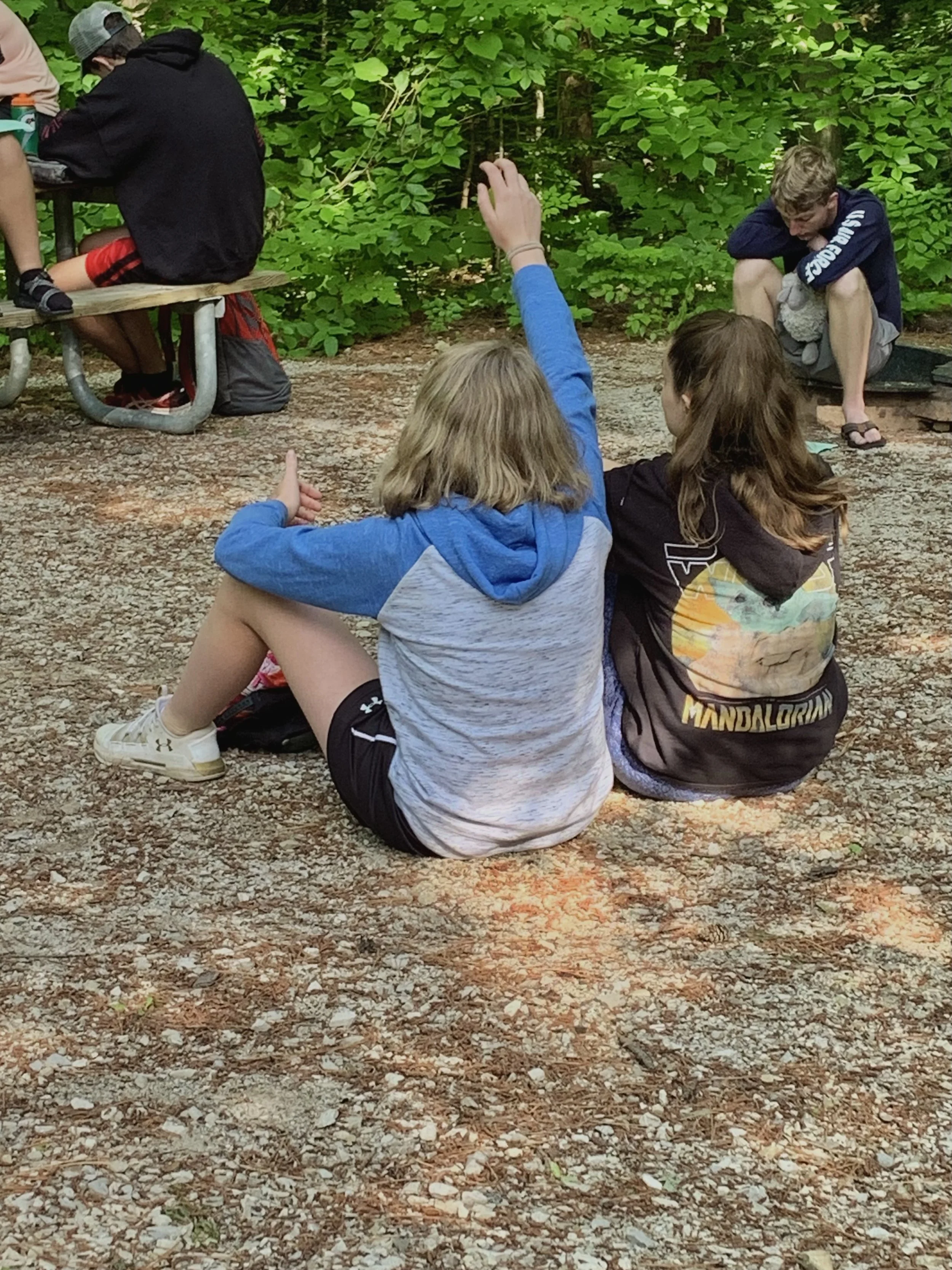 Two girls sitting on the ground in a wooded area, one with a raised hand, and two boys in the background, one sitting on a bench and the other crouching near the ground.