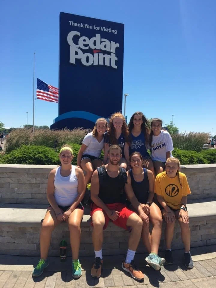 Group of eight young people sitting and standing in front of a sign that says "Thank You for Visiting Cedar Point," with American flag waving nearby on a sunny day.