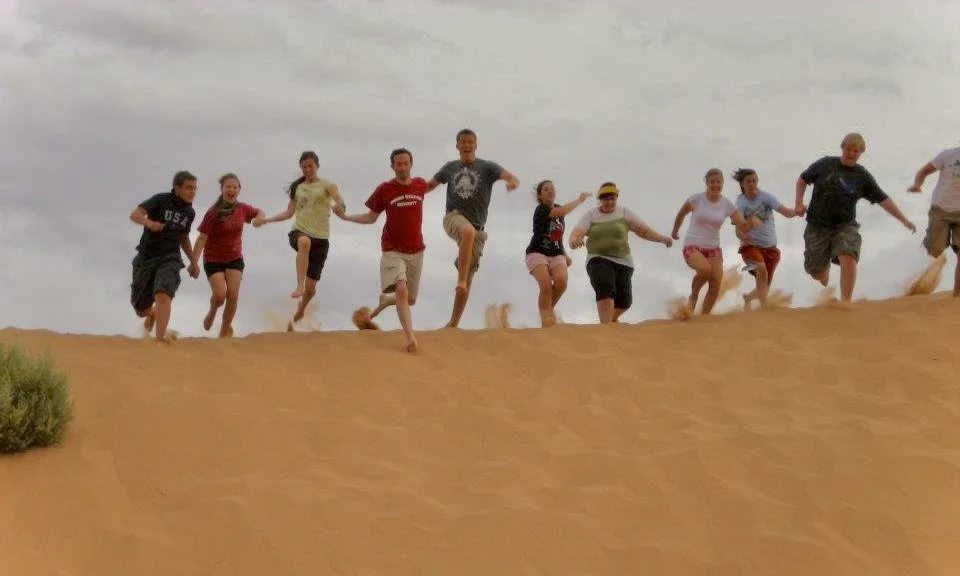 Group of children running and jumping on a sand dune in a desert with cloudy sky behind.