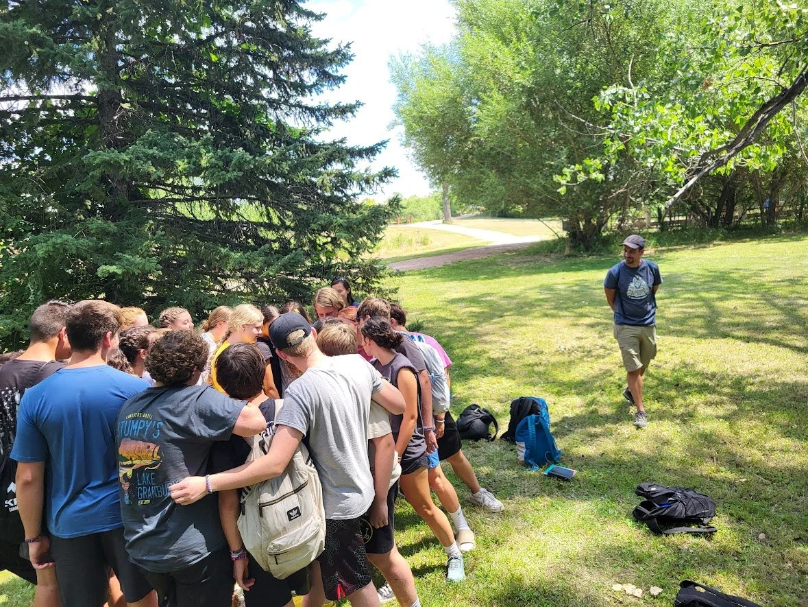 Group of children gathered outdoors, listening to a man giving instructions, with backpacks on the ground, in a grassy area with trees.