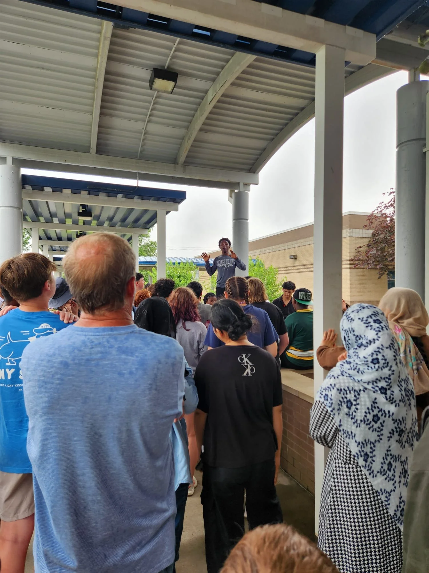 A diverse group of people gathered outdoors under a covered walkway, listening to a speaker standing on an elevated platform gesturing with hands.