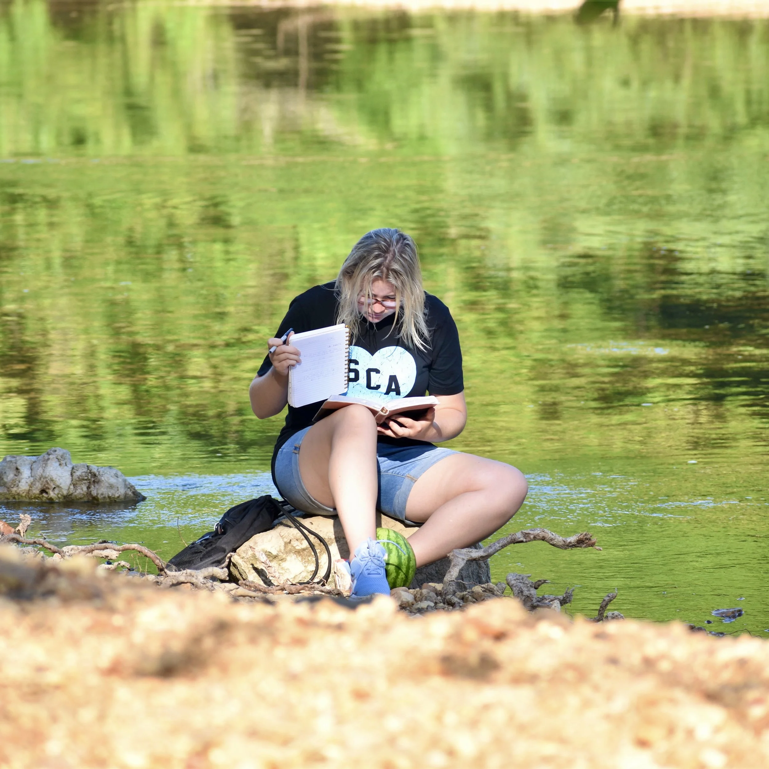 A young woman sitting on rocks by a body of water, reading a book with a notebook in hand, surrounded by nature.