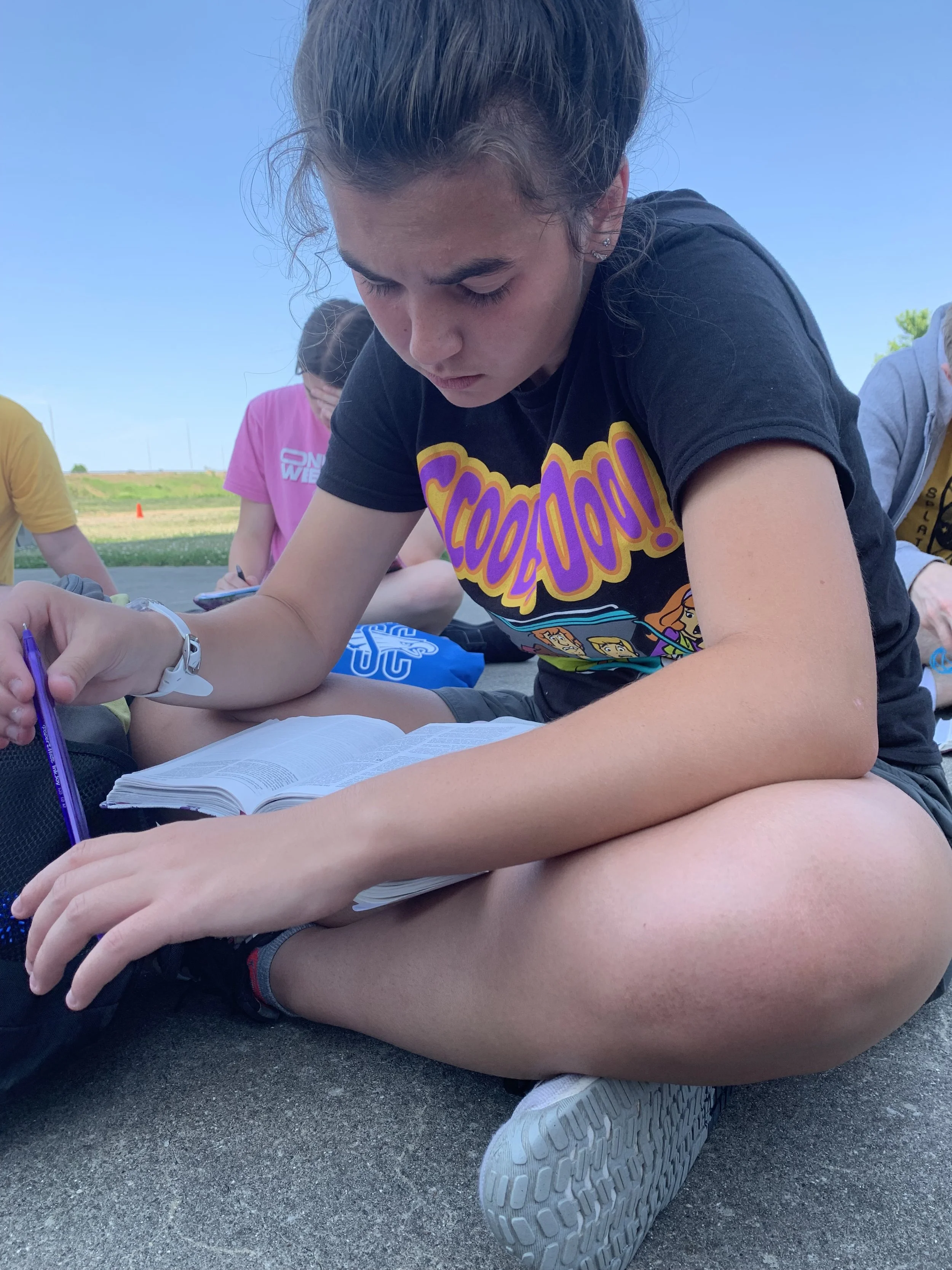 A girl sitting outside on the ground, reading from a book with other children in the background.