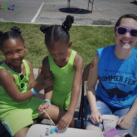 Three young girls sitting at a table outdoors, smiling. Two girls are wearing bright green dresses, and the third girl is wearing sunglasses and a blue T-shirt that says 'Summer Fest.' The girls appear to be at a summer event or festival.