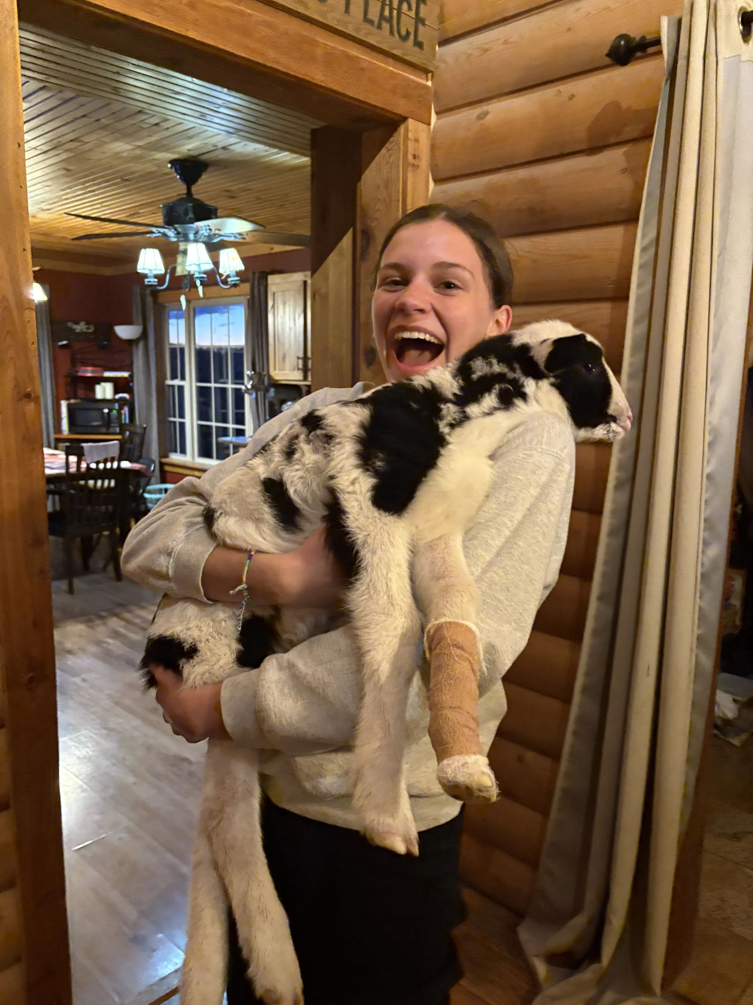A young woman holding a black and white puppy inside a wooden house, smiling brightly.