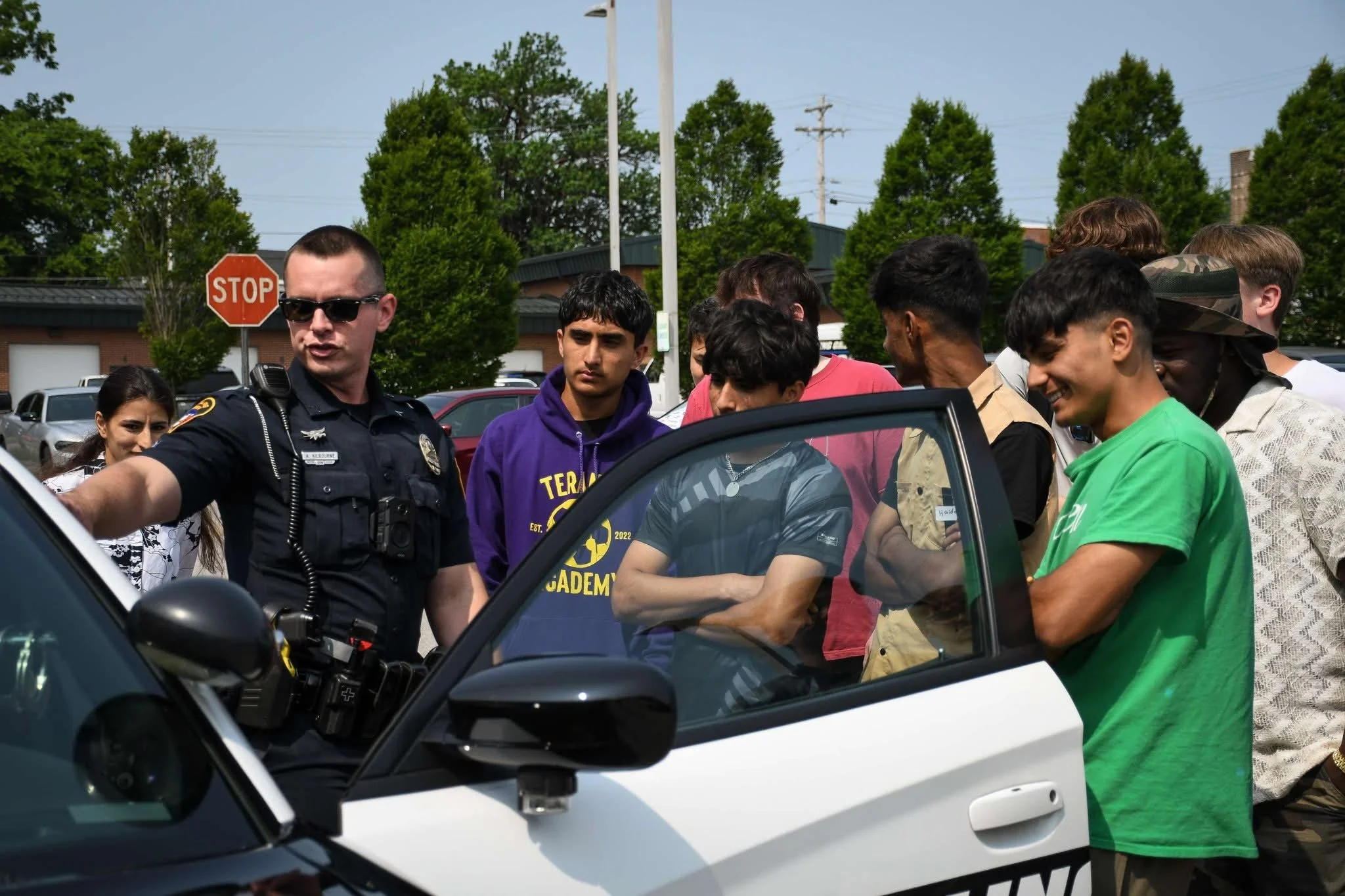 A police officer and a group of diverse young people gathered around a police car, engaging in a discussion outdoors on a sunny day.