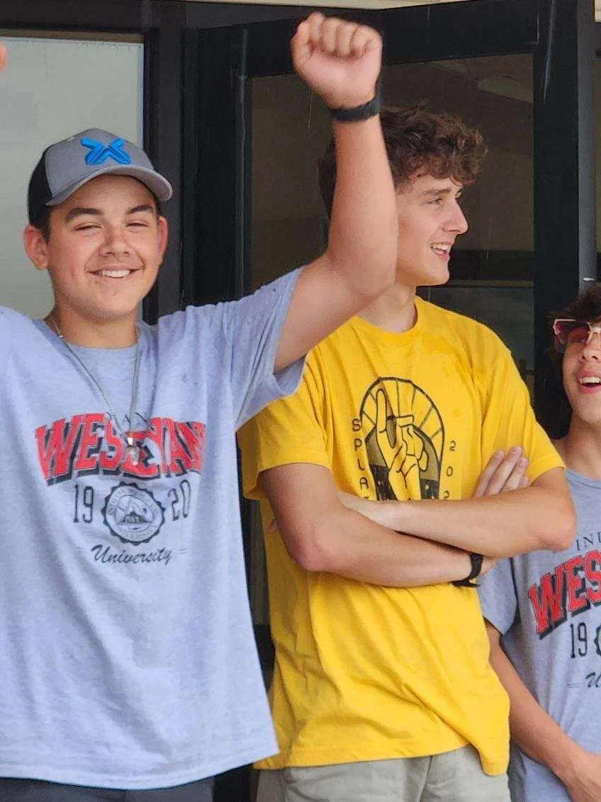 Three teenagers standing outside, smiling and interacting, with two of them wearing university t-shirts and one with their arm raised in a celebratory gesture.