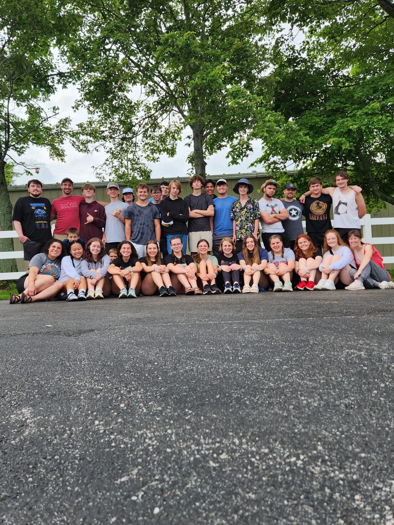 A large group of teenagers and young adults posing outdoors in front of a white fence with green trees behind them. The group is smiling, with some sitting on the ground and others standing behind.