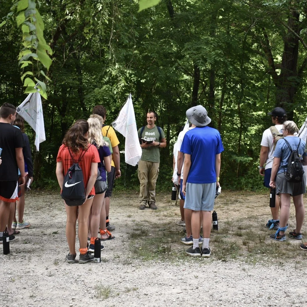 Group of people on a nature walk or outdoor class in a wooded area, listening to a guide or instructor.