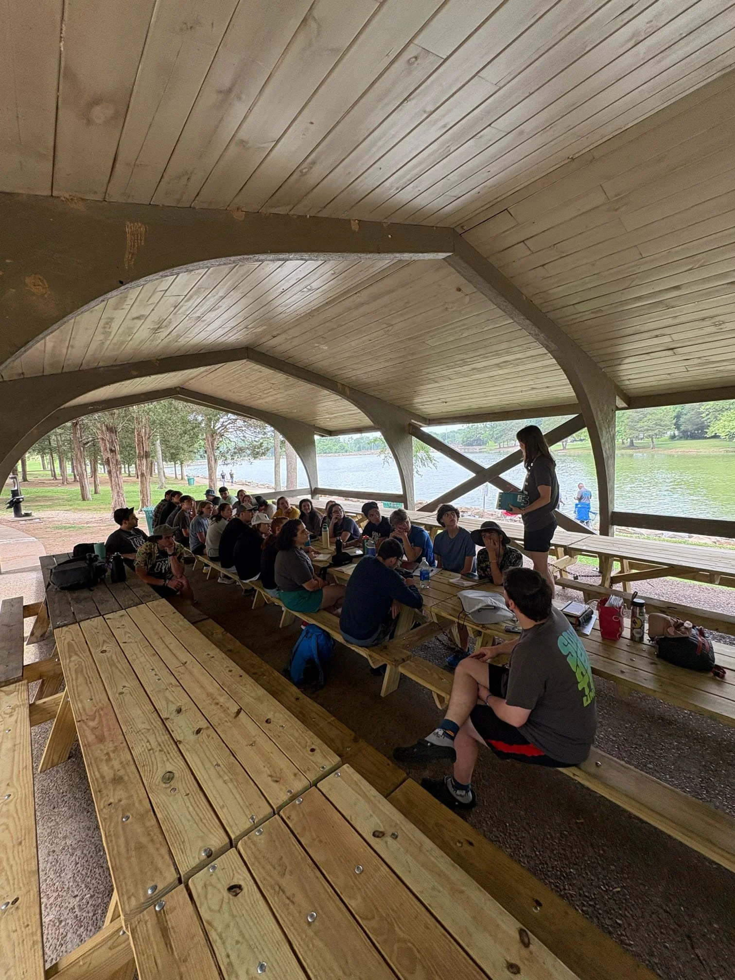 A group of students sitting at picnic tables under a shelter near a lake, with a teacher standing and speaking to them.