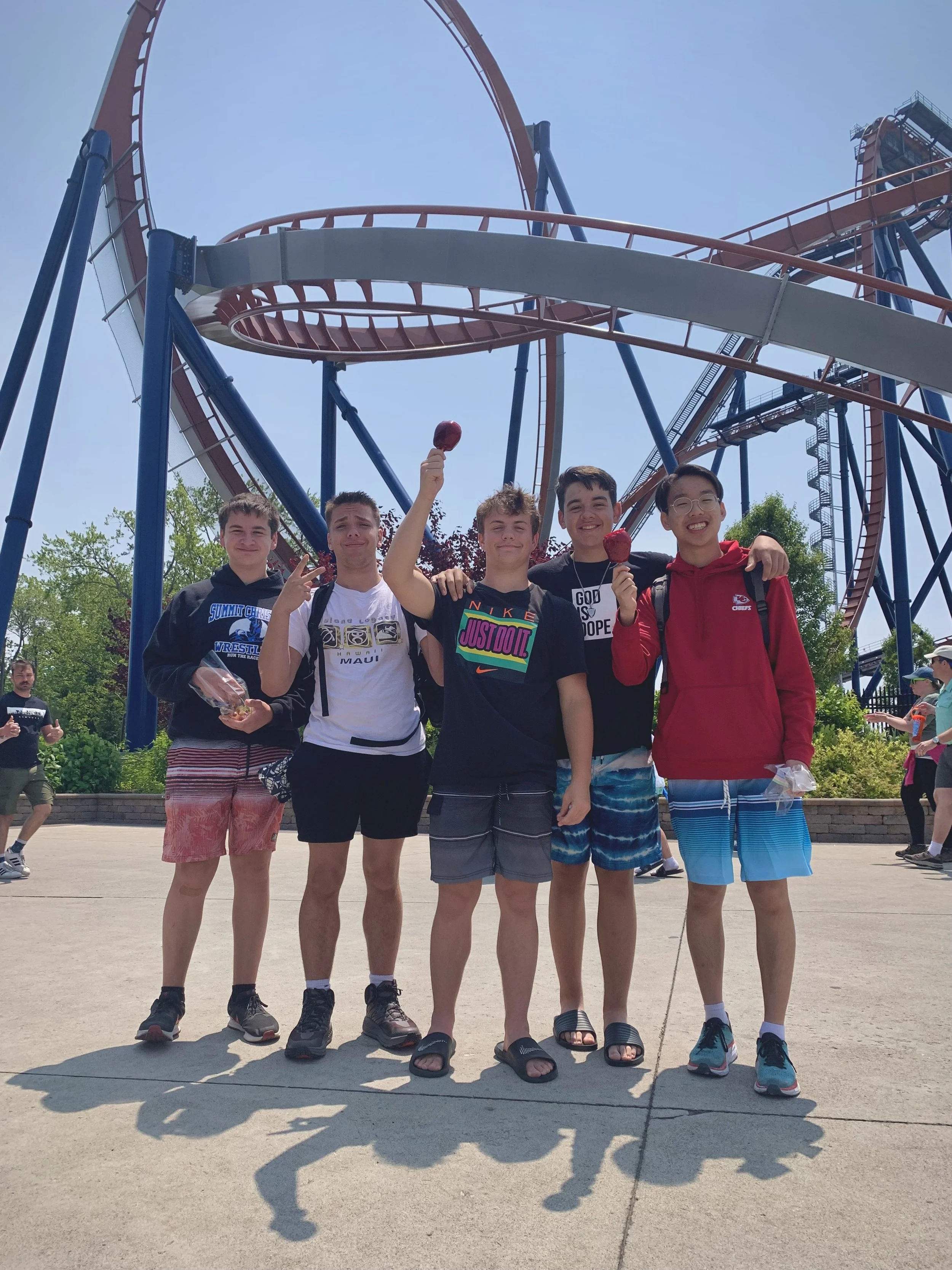 Five teenage boys standing in front of a roller coaster, smiling and holding snacks and ice cream on a sunny day at an amusement park.