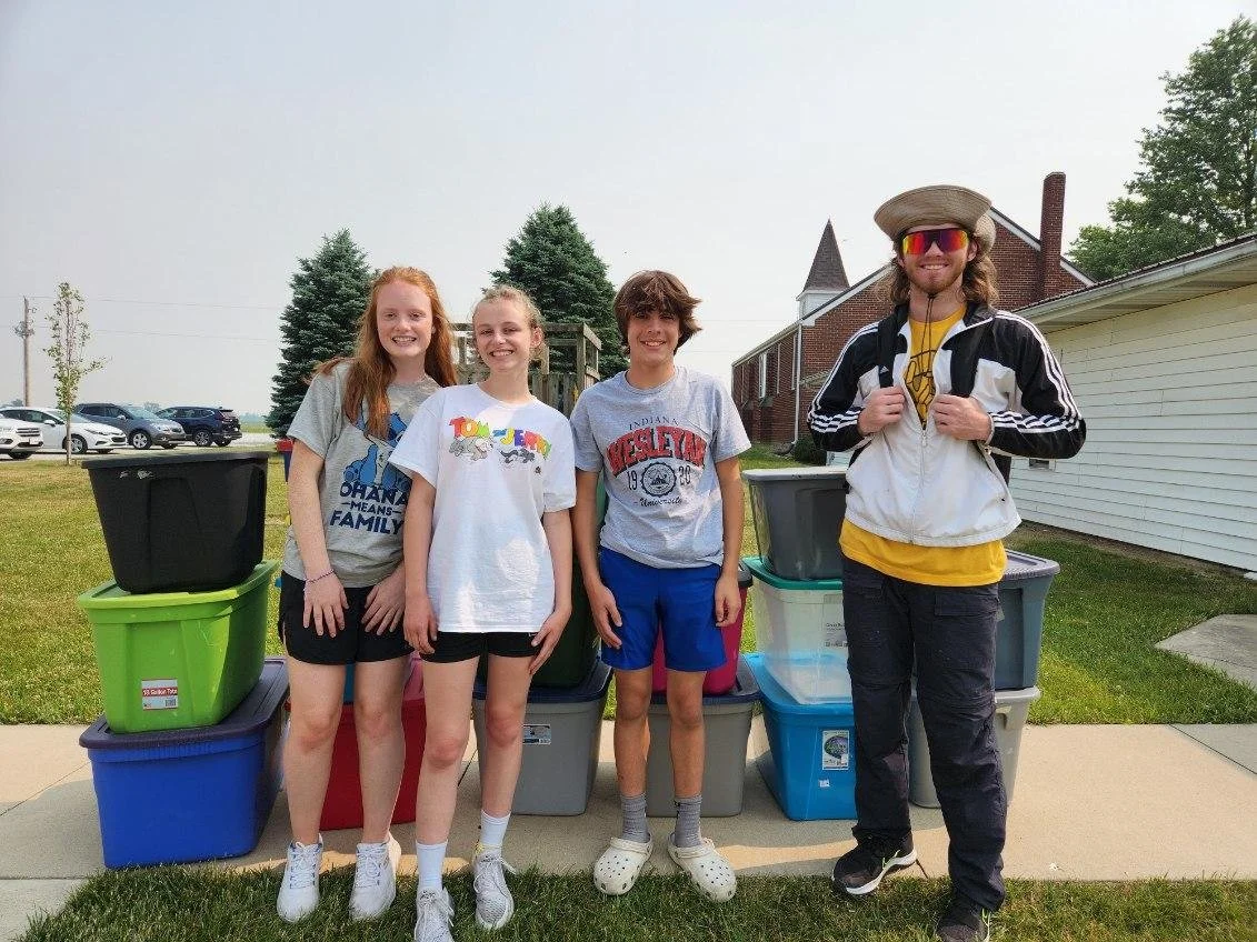 Four children standing outdoors in front of colorful storage bins, smiling at the camera, with a man on the far right wearing sunglasses, a hat, and a jacket.