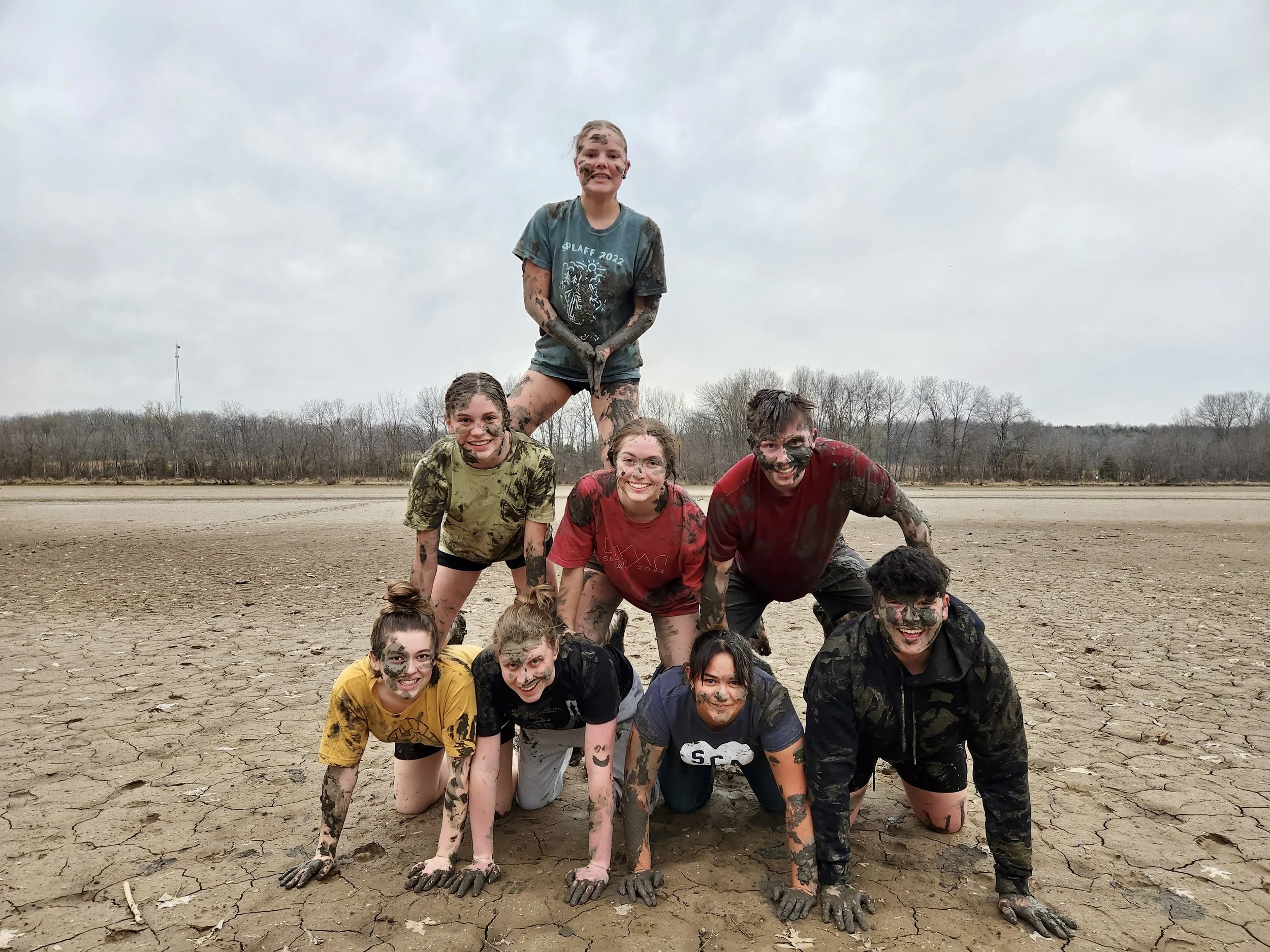 Group of ten people in muddy clothes posing in a human pyramid on cracked, dry ground outdoors with a cloudy sky and distant trees in the background.