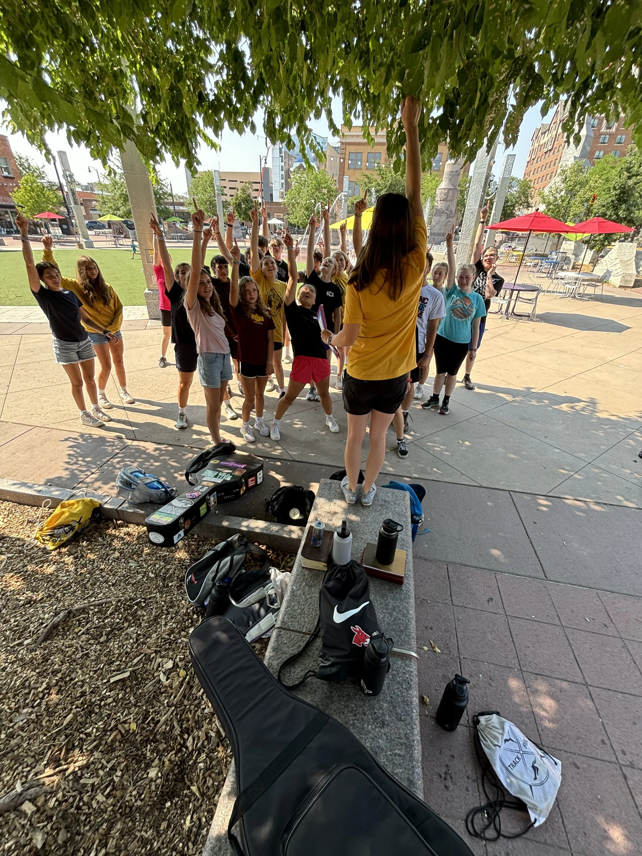 Group of young girls participating in outdoor fitness or exercise class, led by an instructor wearing a yellow shirt, under the shade of a large leafy tree.