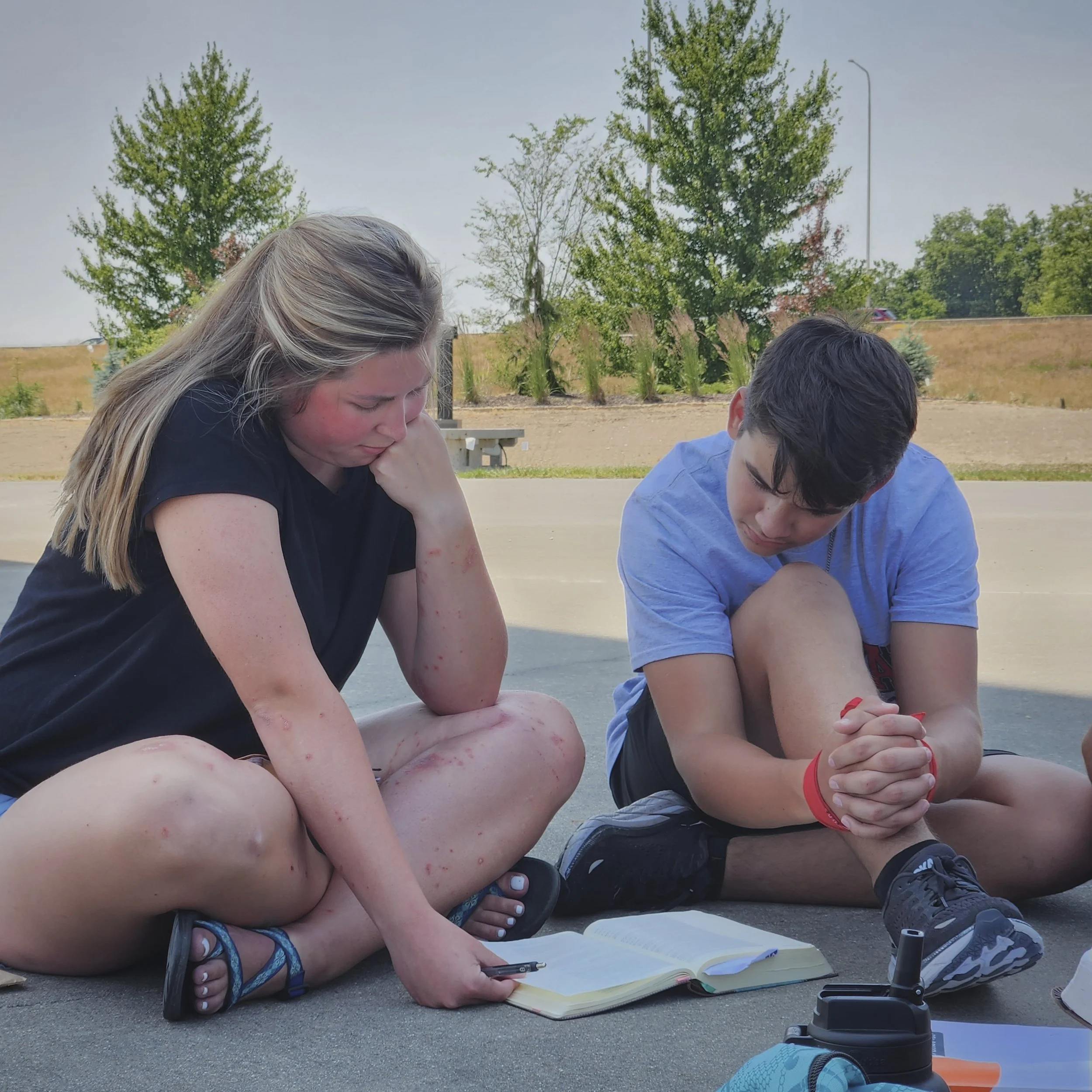 Two teenagers sitting cross-legged on a basketball court, reading a book together with a notebook and supplies nearby, outdoors on a sunny day with trees in the background.