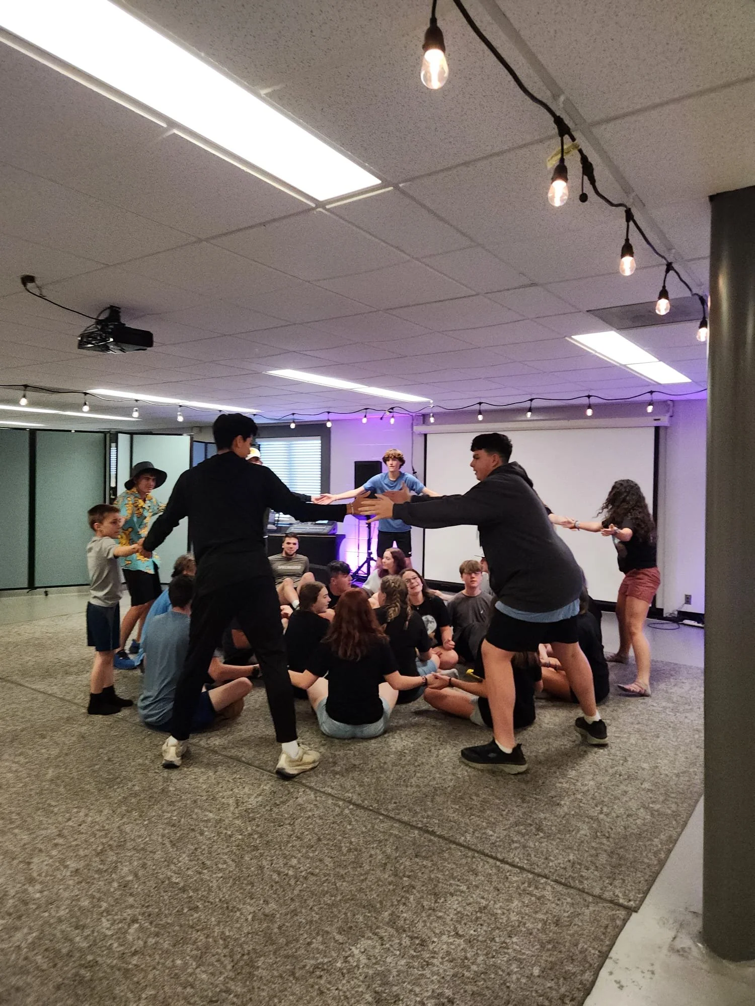 Group of young people sitting on the floor in a room, while others are holding hands and forming a circle around them, in a casual setting with string lights and a projector in the background.