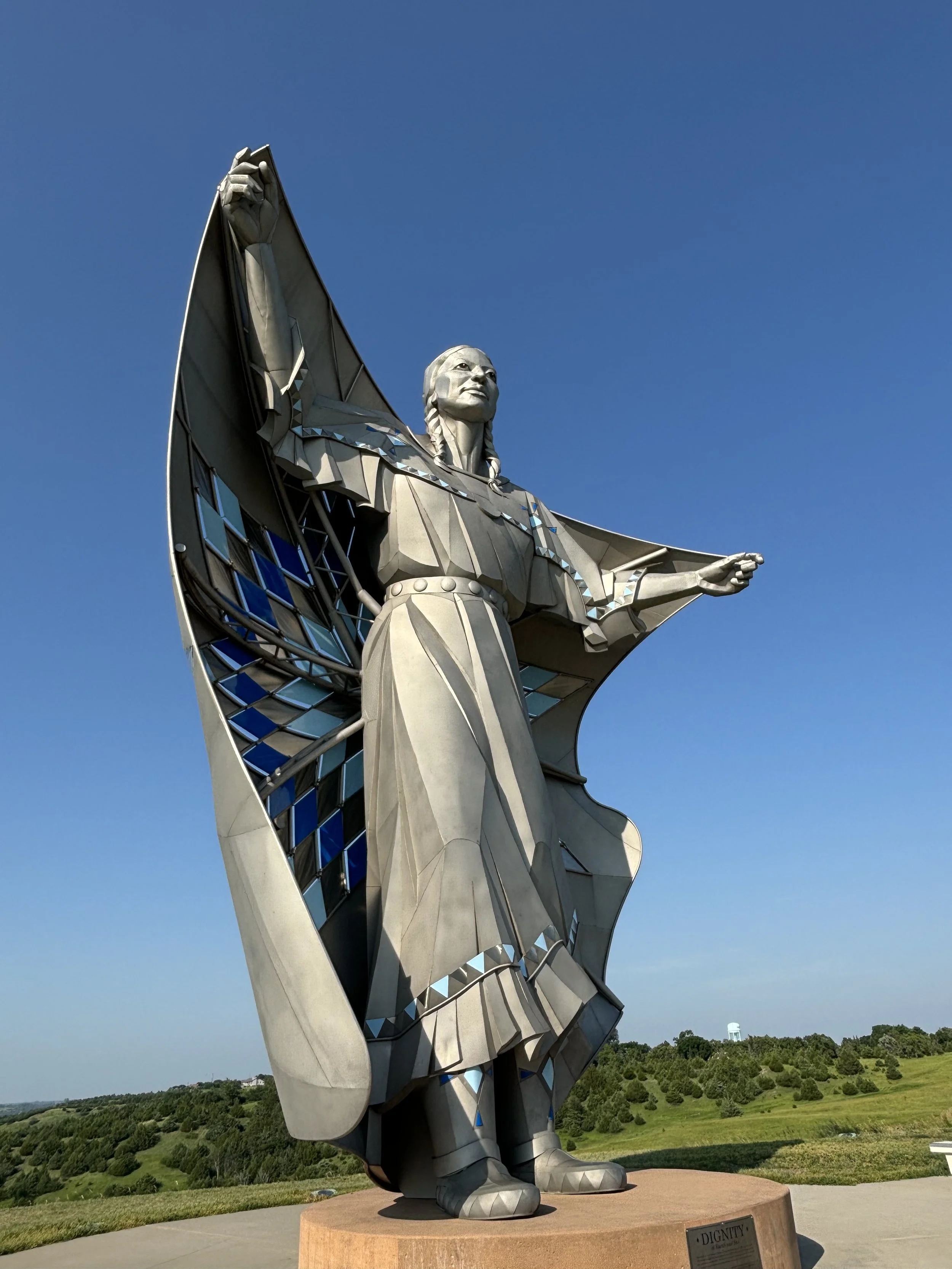 Large statue of a Native American man with arms outstretched, standing outdoors with green hills and a clear blue sky in the background.