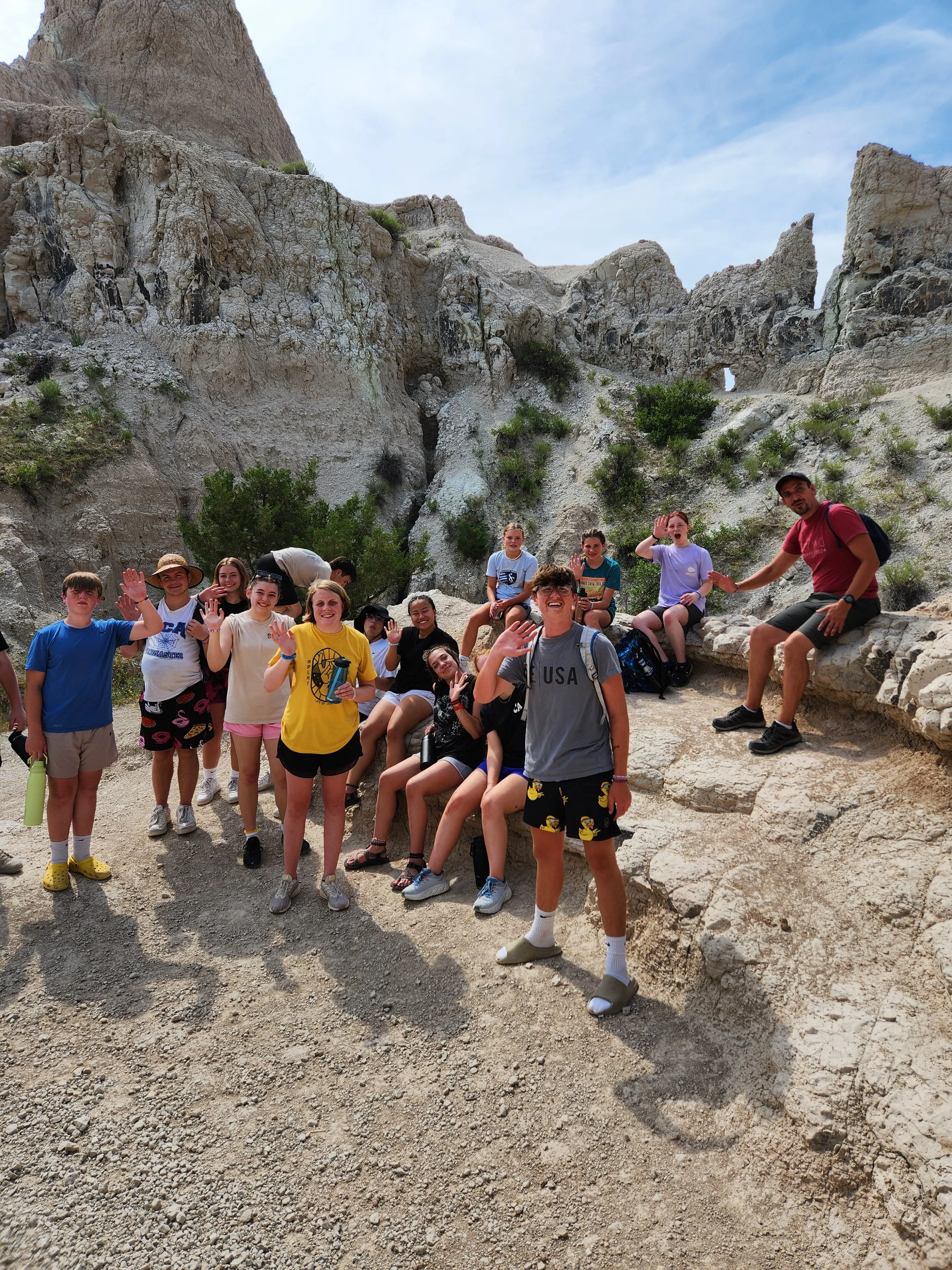 A group of children and one adult adventure hiking in a rocky, desert landscape with rugged cliffs and sparse vegetation, smiling and waving at the camera.