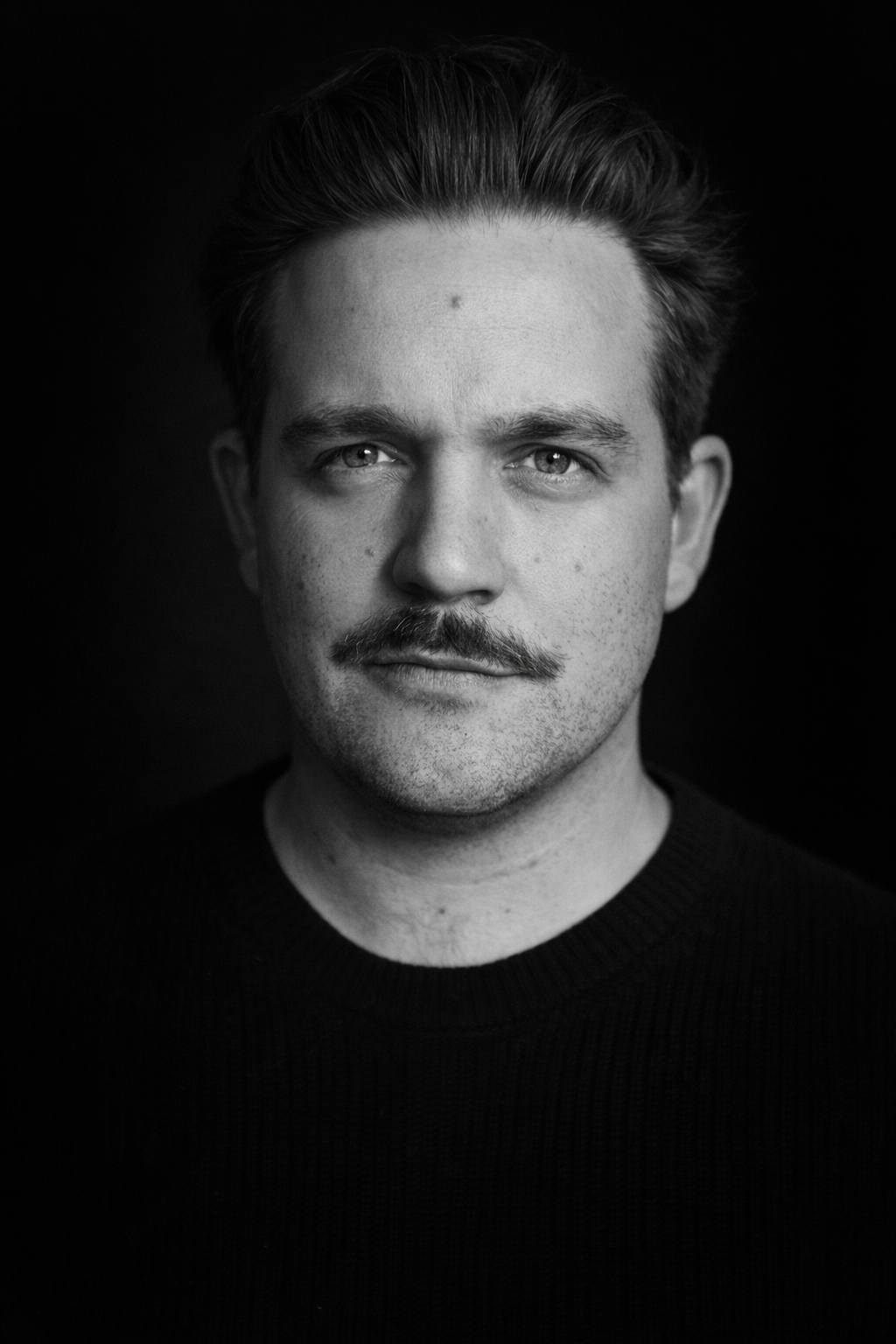 Black and white portrait of a man with a mustache, short hair, and a serious expression, wearing a dark knit sweater against a dark background.