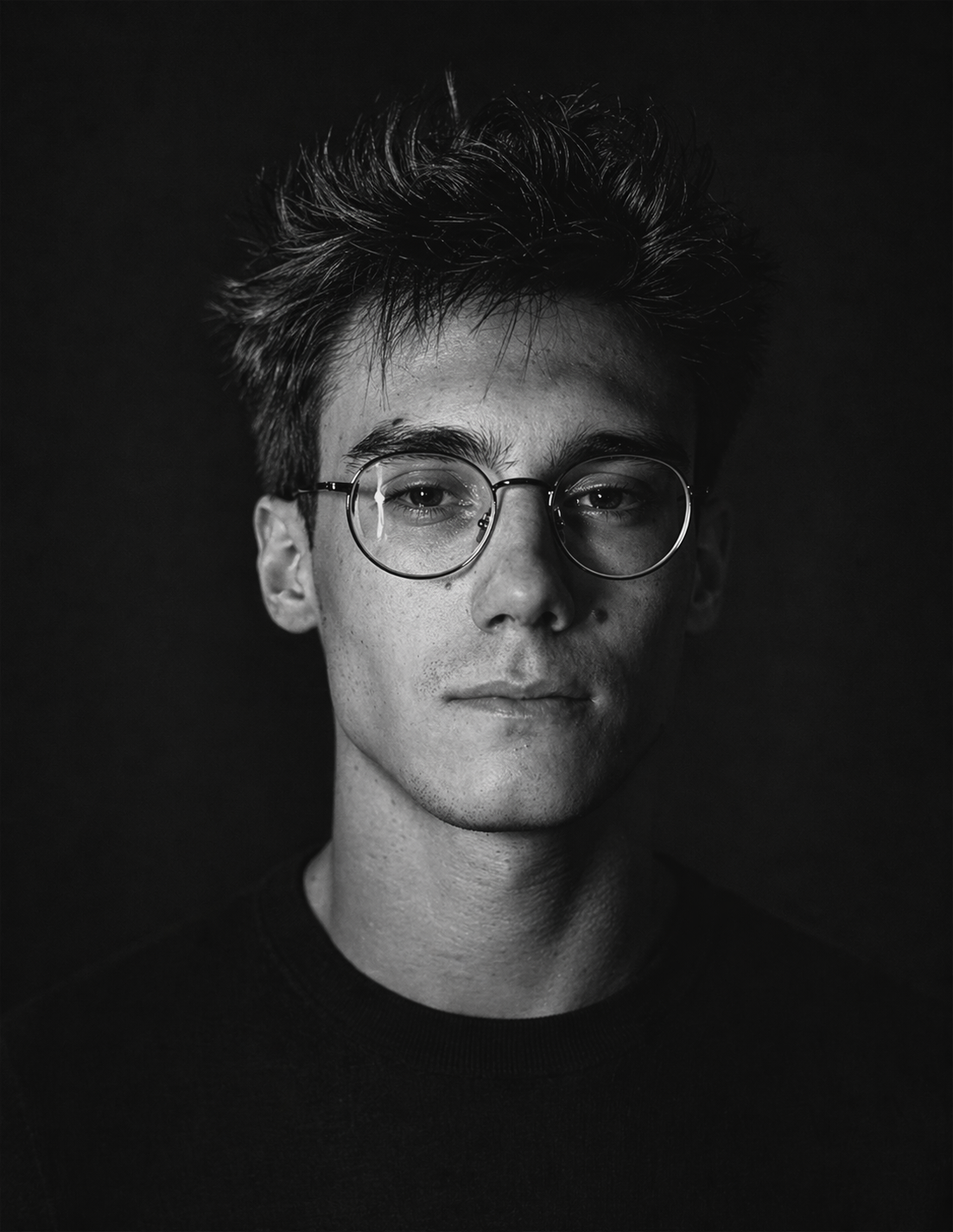 Black and white portrait of a young man with glasses, short messy hair, and a neutral expression, against a dark background.