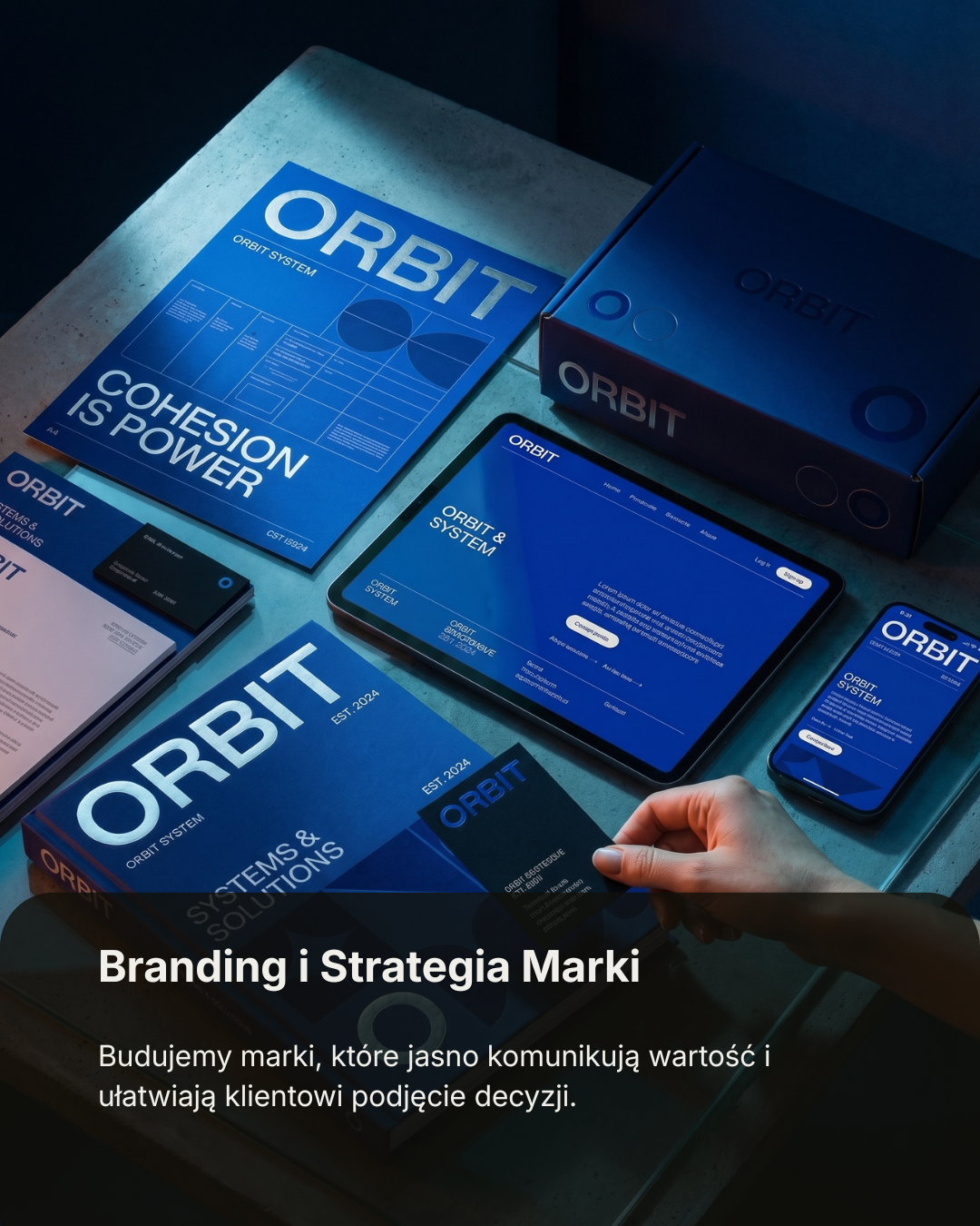 A workspace with branding materials for a company called Orbit, including a large book, a tablet, a smartphone, and a box, all in blue themed with the Orbit logo, with a hand holding the book and a Polish caption about branding and strategy.
