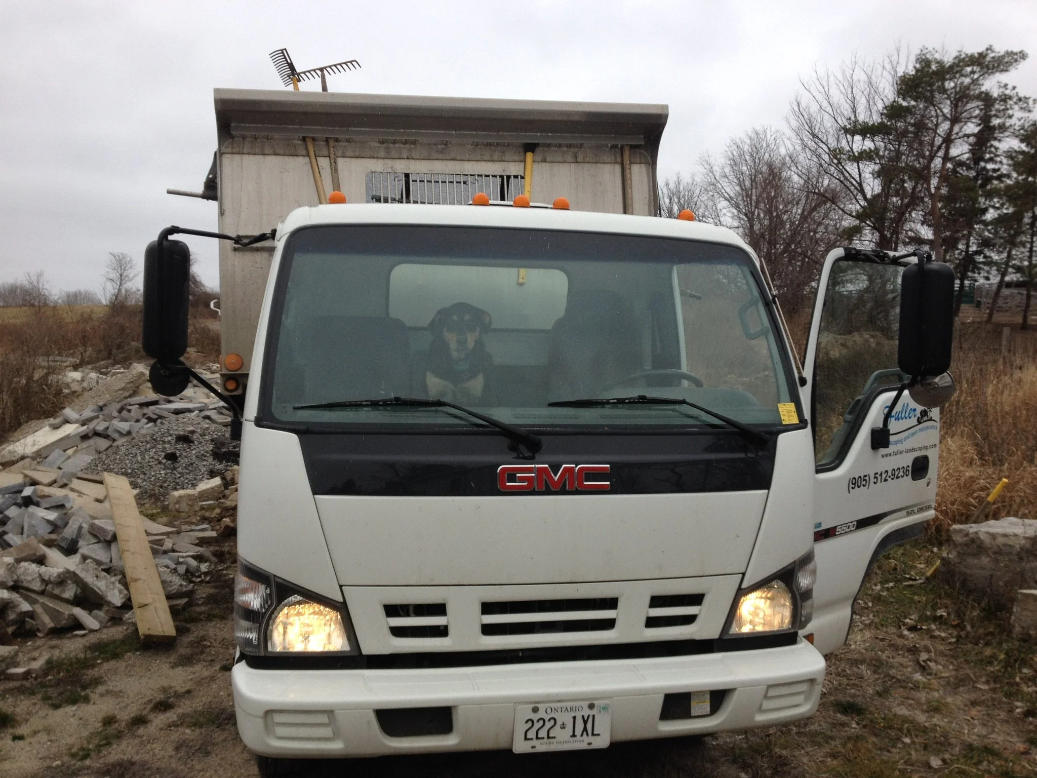 A white GMC delivery truck with a dog sitting inside behind the windshield. The truck is parked outdoors on a construction or gravel lot, with a pile of rocks and debris visible to the side. The truck's door is open and the vehicle has an Ontario license plate.